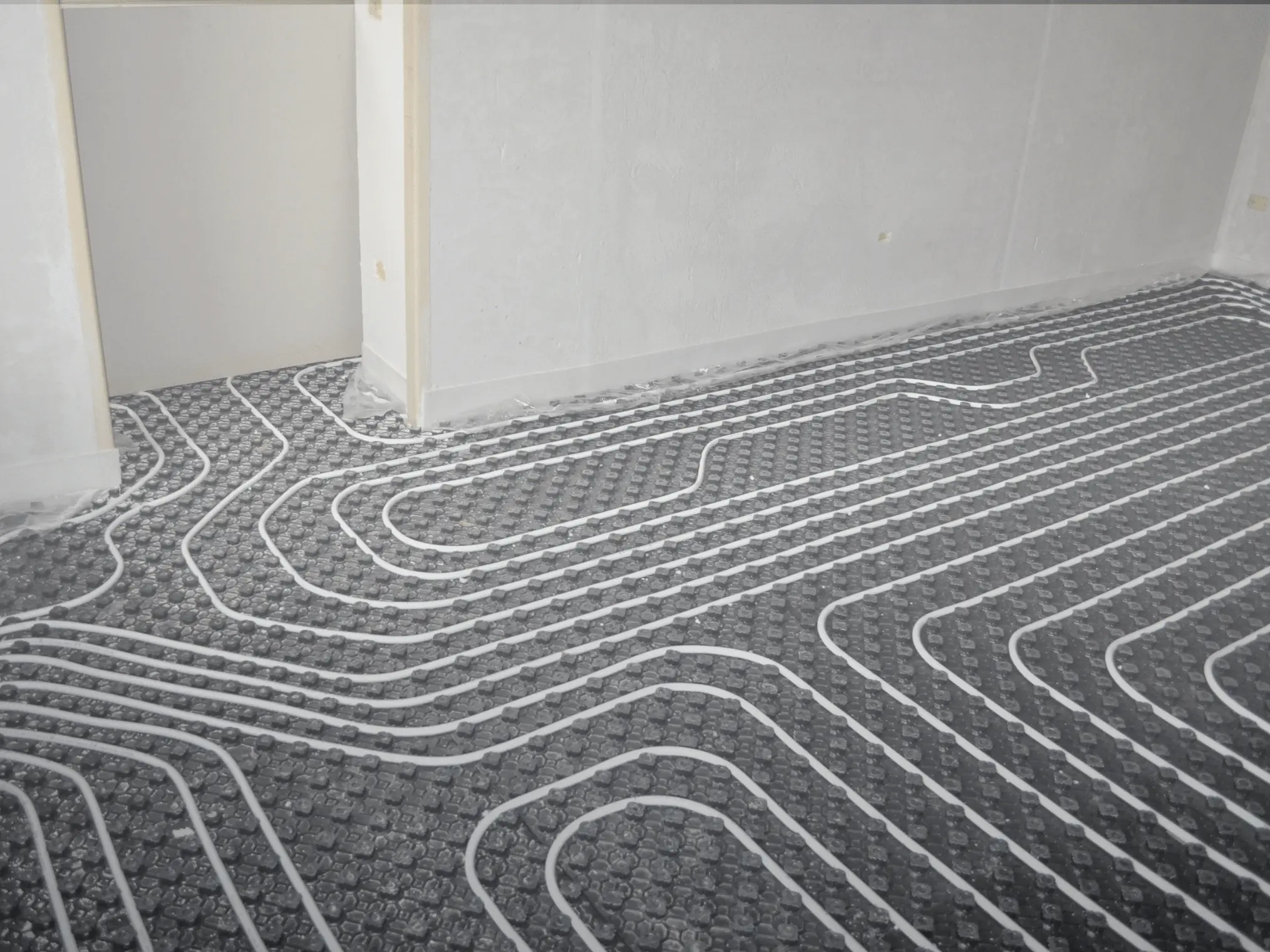 Underfloor heating system installation with visible coiled pipes on a textured black surface in a room with unfinished walls.