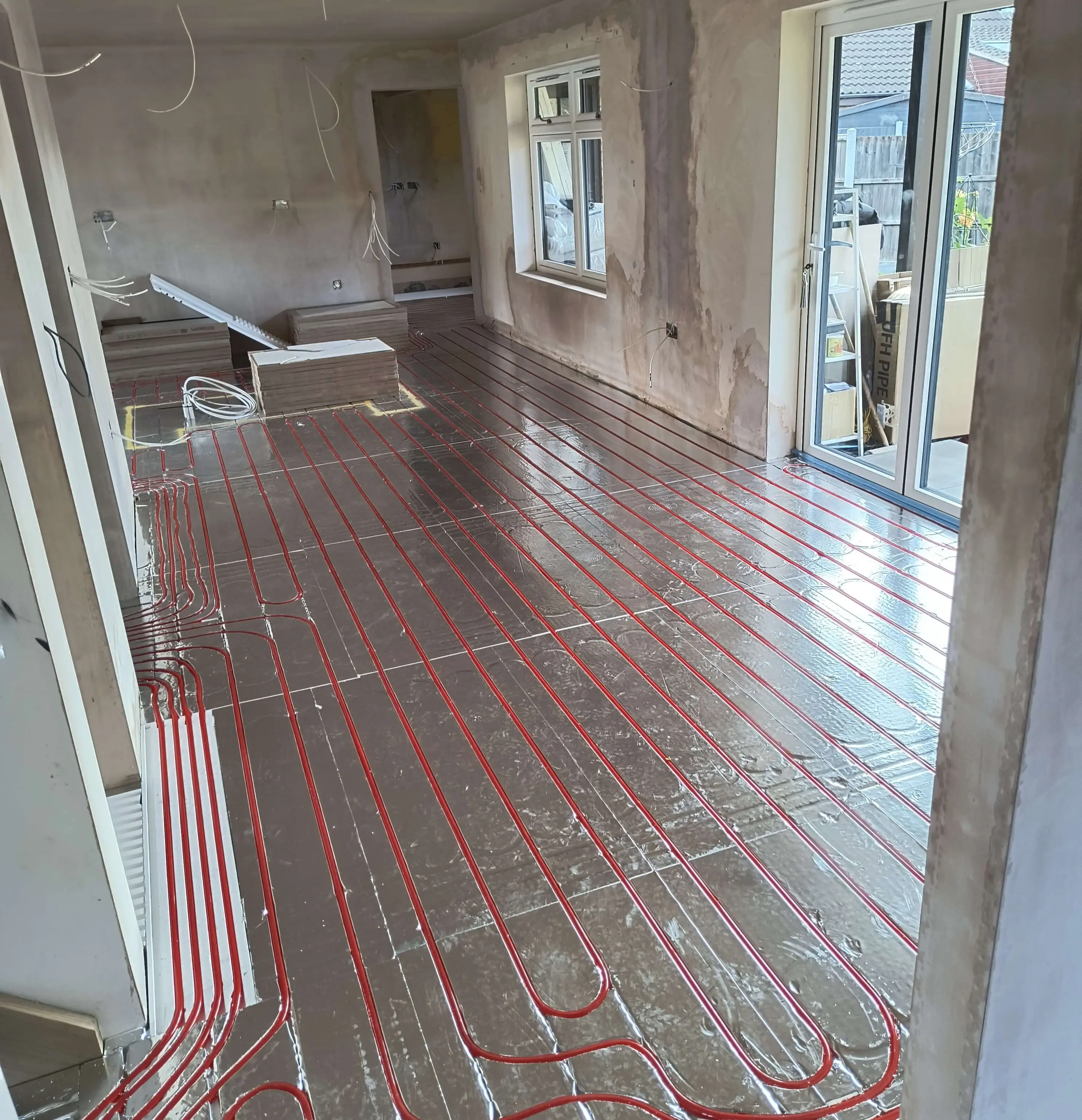 Room under renovation with exposed underfloor heating pipes laid over reflective insulation. Walls are unfinished, with windows and glass doors.