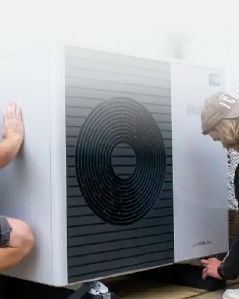 Two workers install a large outdoor heat pump unit against a white wall, focusing on securing it into place, with tools nearby.