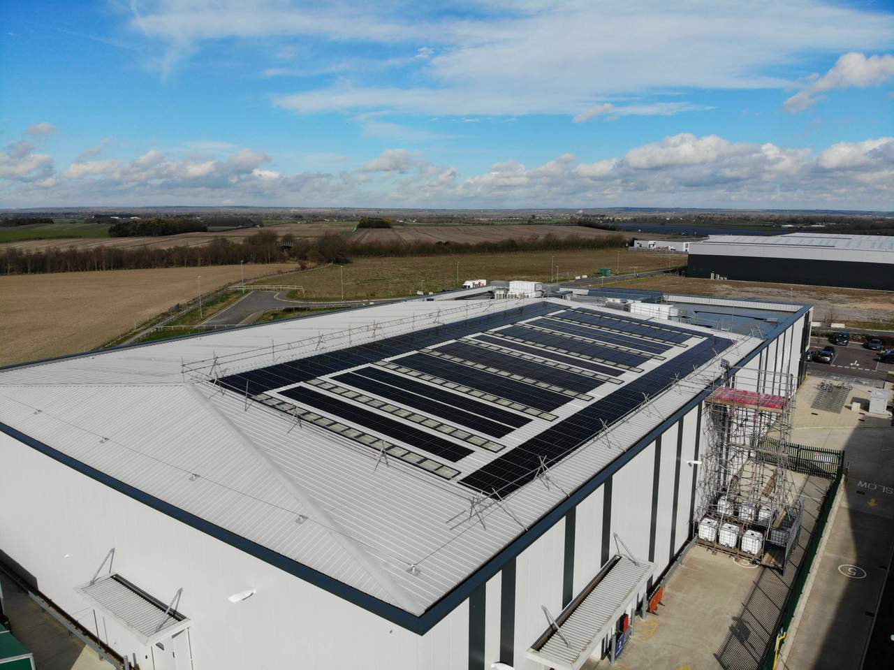 Aerial view of a large industrial building with solar panels on the roof, surrounded by fields and a clear blue sky.