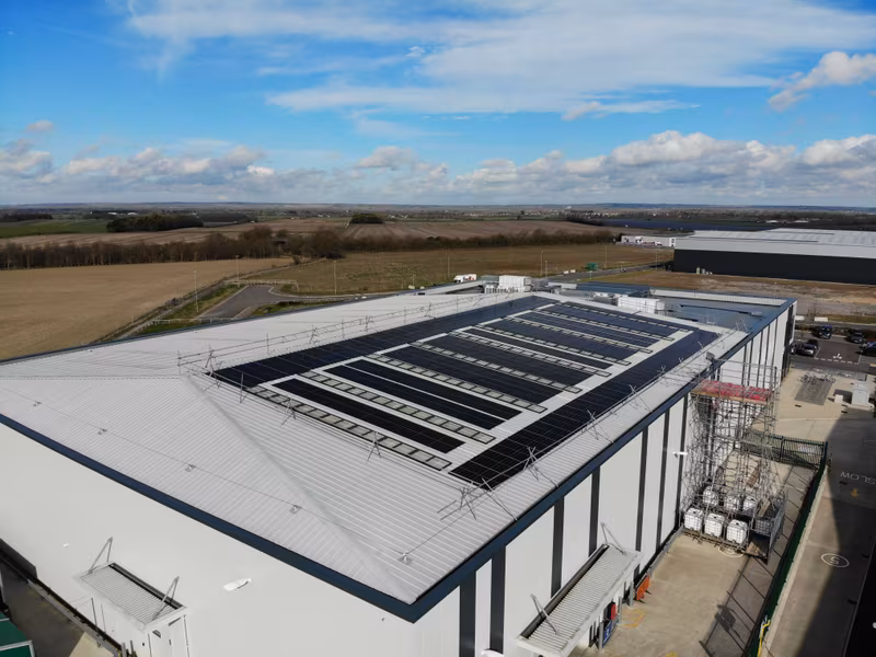 Aerial view of a large industrial building with solar panels on the roof, surrounded by fields and a clear blue sky.