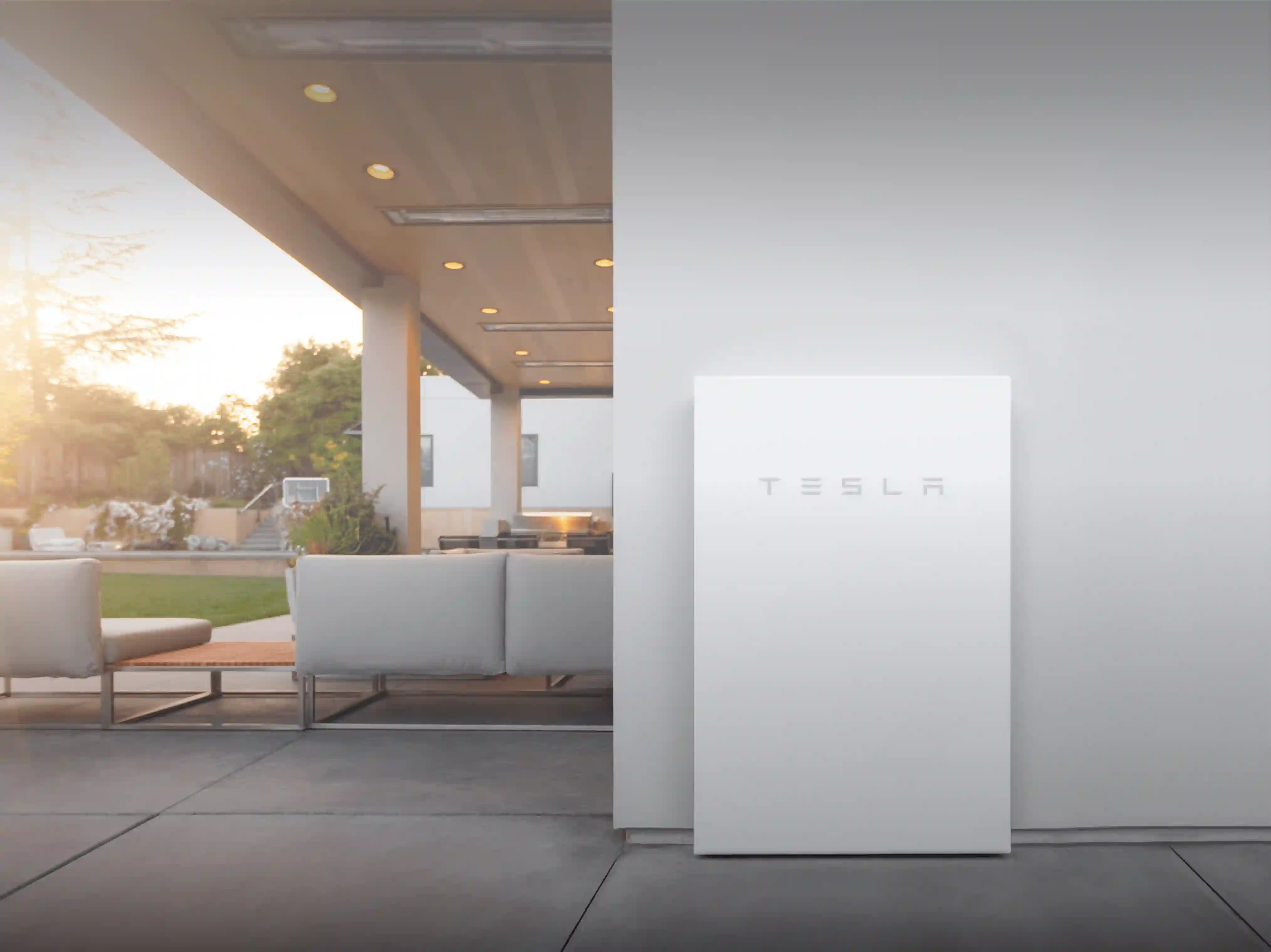 A white Tesla Powerwall installed on an exterior wall of a modern house with an open patio and outdoor seating area.