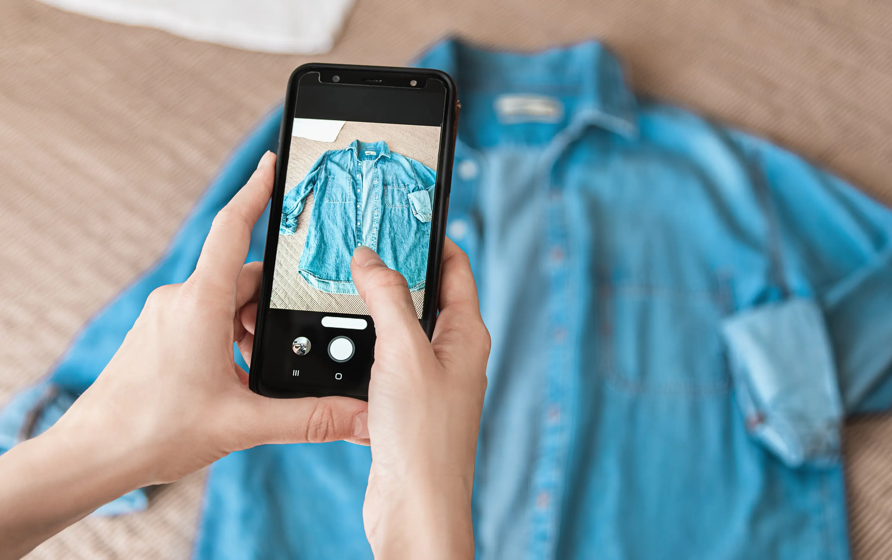 Person photographing a blue denim shirt with a smartphone, focusing on the garment's details.
