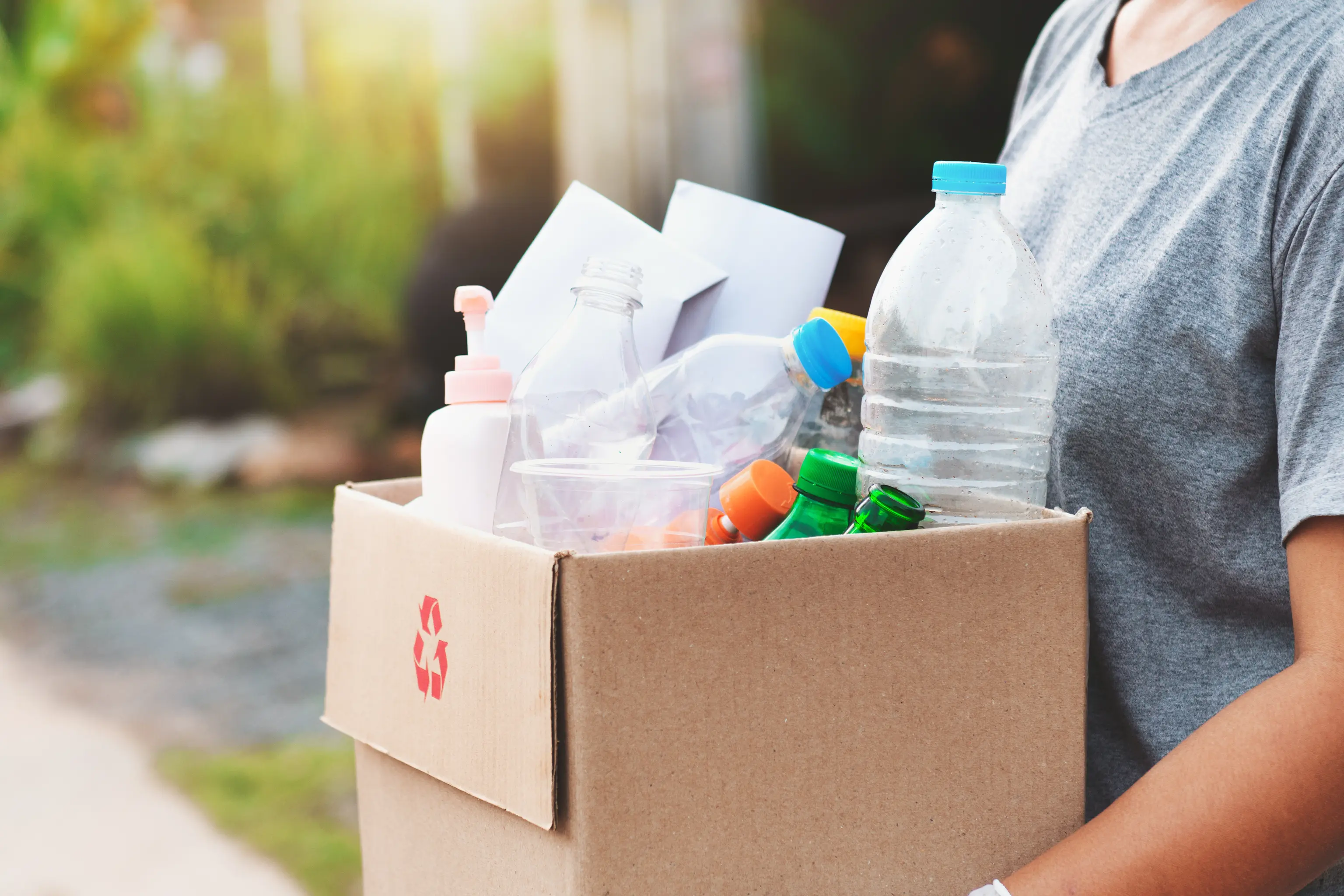 Person holding a cardboard box filled with plastic bottles and containers for recycling, outdoors in a sunny setting.