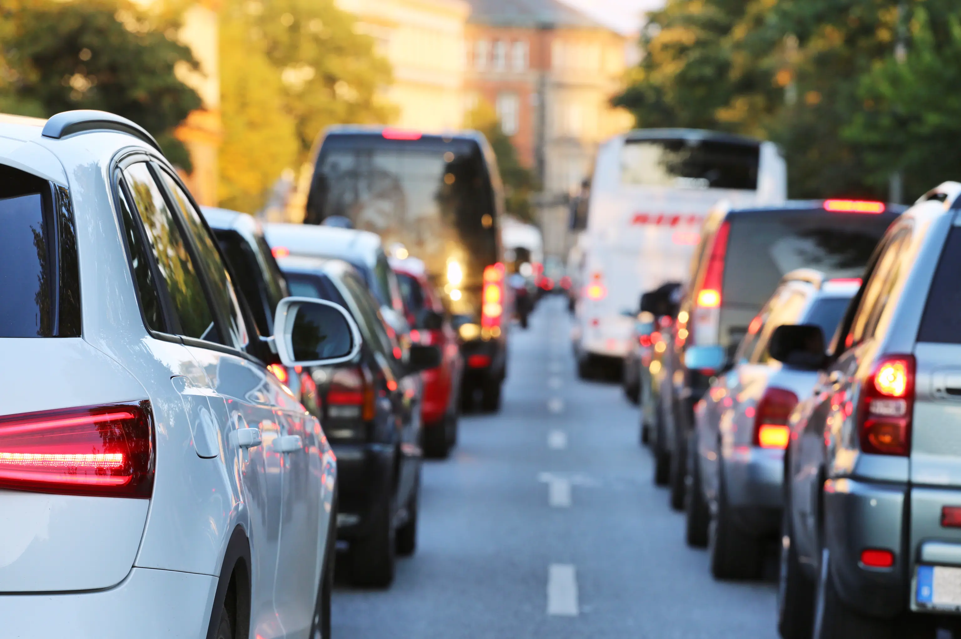 Traffic jam on a city street with cars and buses lined up closely under a clear sky, surrounded by trees and buildings.