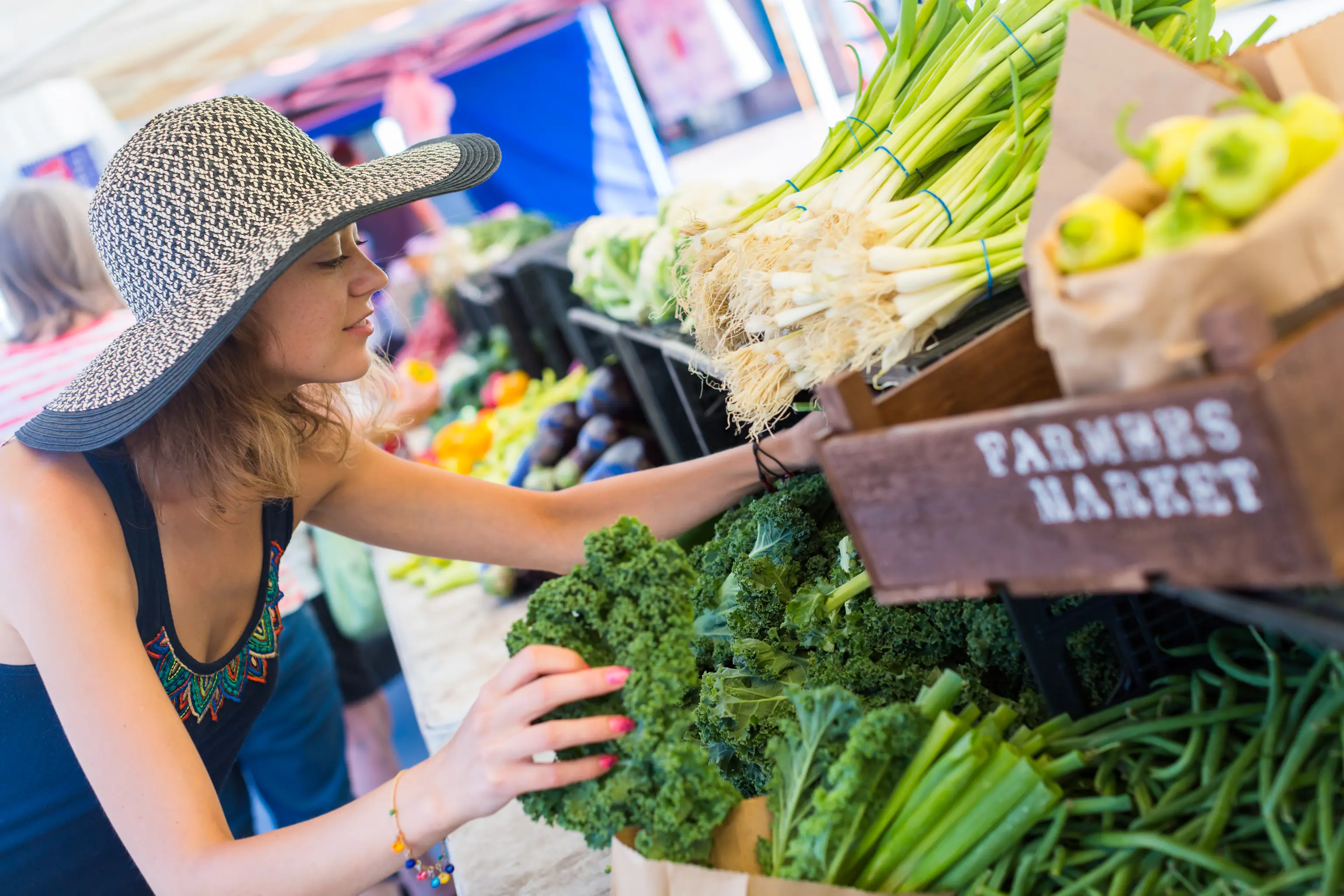 Woman in a sun hat selecting fresh vegetables at a farmers market stall, surrounded by greens, peppers, and colorful produce.