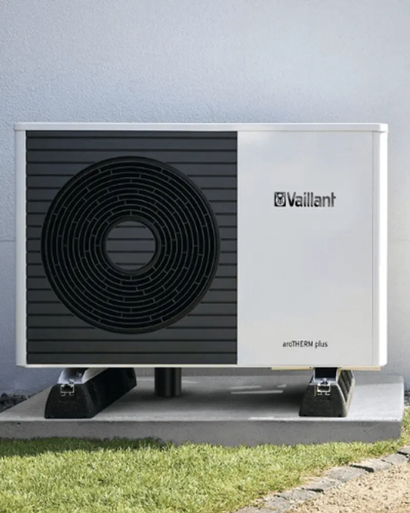 Outdoor heat pump unit mounted on a concrete slab, featuring a large circular vent and the Vaillant logo on a white and black casing.
