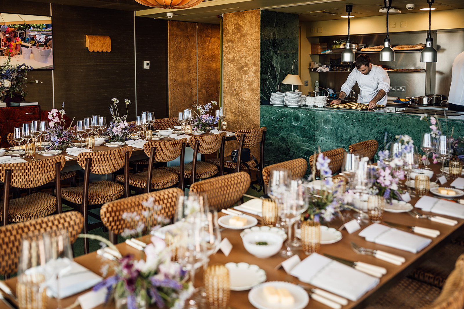 Elegant dining setup with woven chairs, floral centerpieces, and neatly arranged tableware; chef working in the background near a green counter.