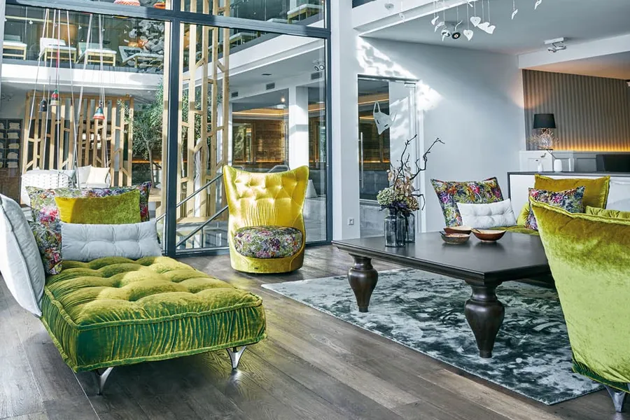 Modern living room with high ceilings, green velvet furniture, floral cushions, a large glass wall, and a cascading white paper chandelier.