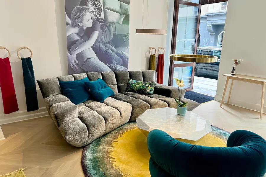 Bretz Showroom mit dem Cloud 7 Sofa im Bezug silberschimmer, davor liegt ein runder blau-gelber Teppich, der farblich zu den Kissen auf dem Sofa passt.