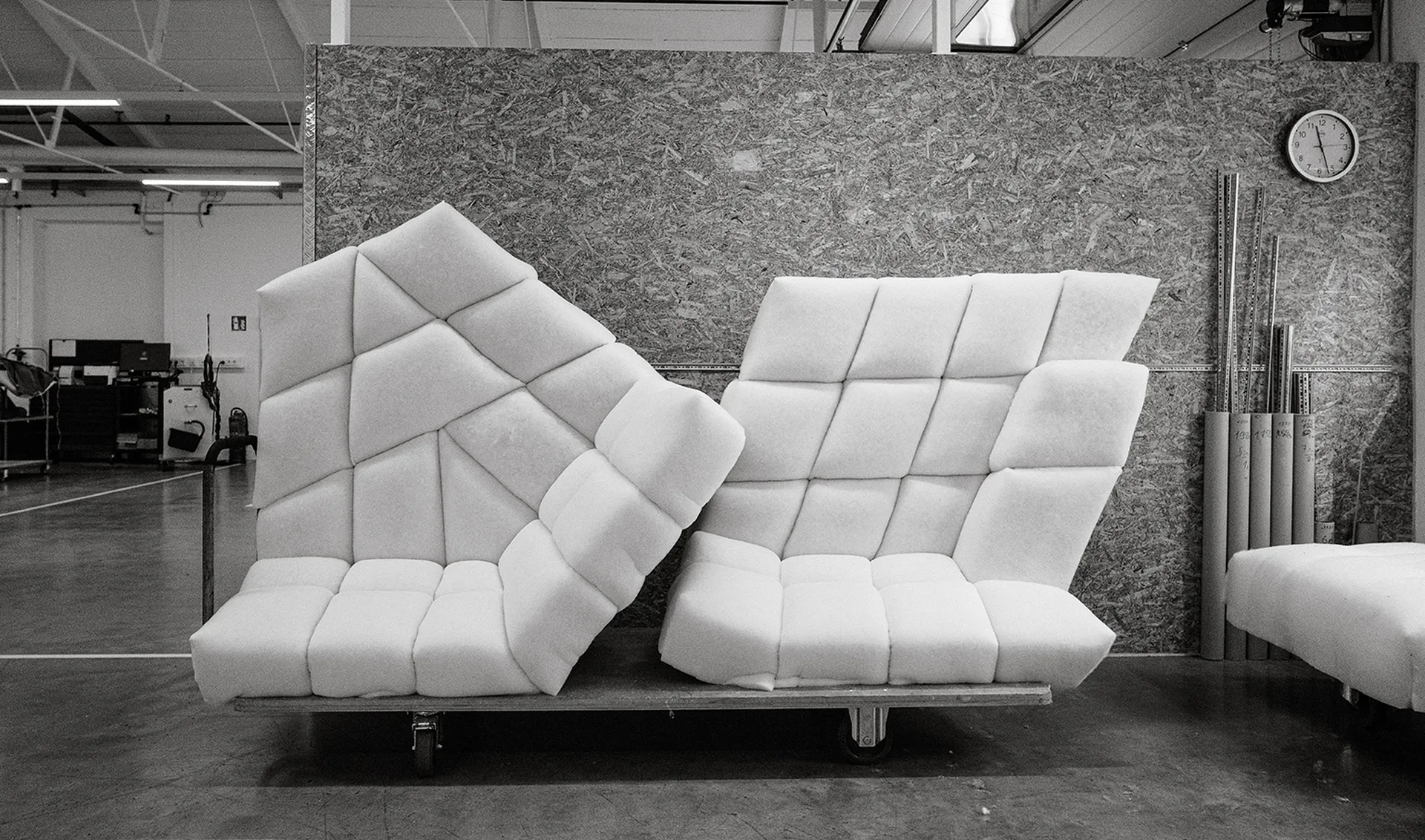 Two modern, geometric armchairs with a quilted pattern, placed in a workshop setting on a wheeled base, with a clock on the wall.