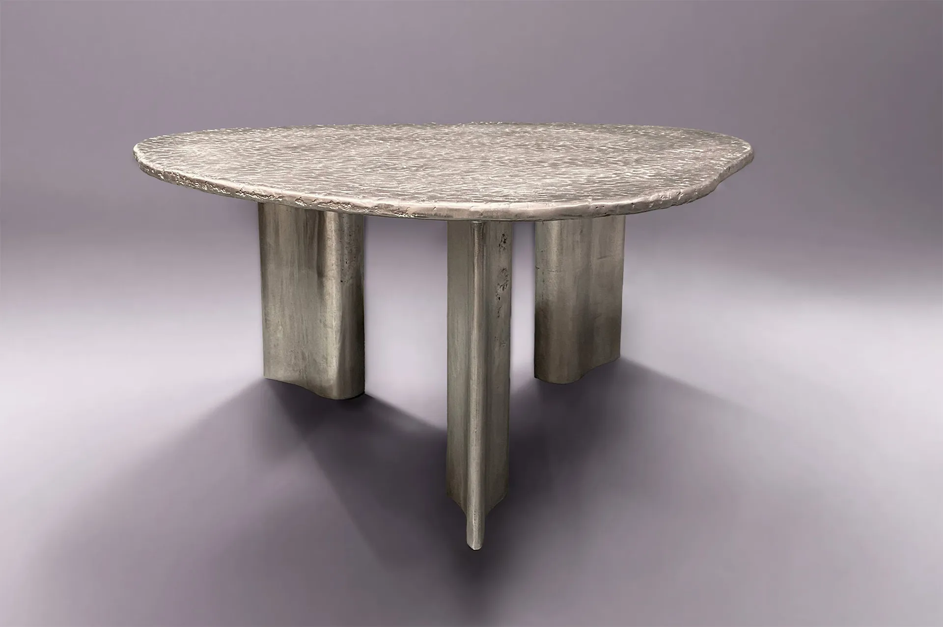 A sculptural table with a textured, irregularly shaped top and three thick, uneven metal legs on a gray background.