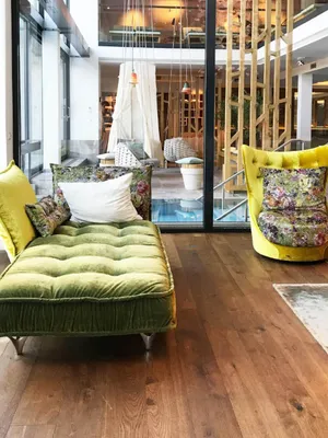 Modern lounge with green velvet chaise and chair, floral cushions, wooden floor, and large windows offering natural light.