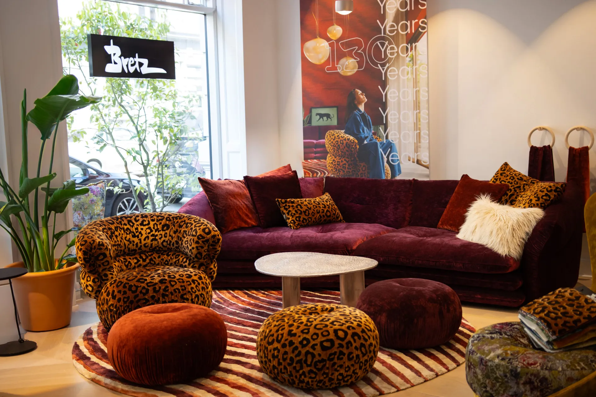 Ein gemütliches Wohnzimmer mit einem dreilagigen dunkelroten Sofa, Sessel mit Leopardenmuster, und einem gestreiften Teppich. Ein großes Fenster und Pflanzen runden die Einrichtung des Showrooms ab.