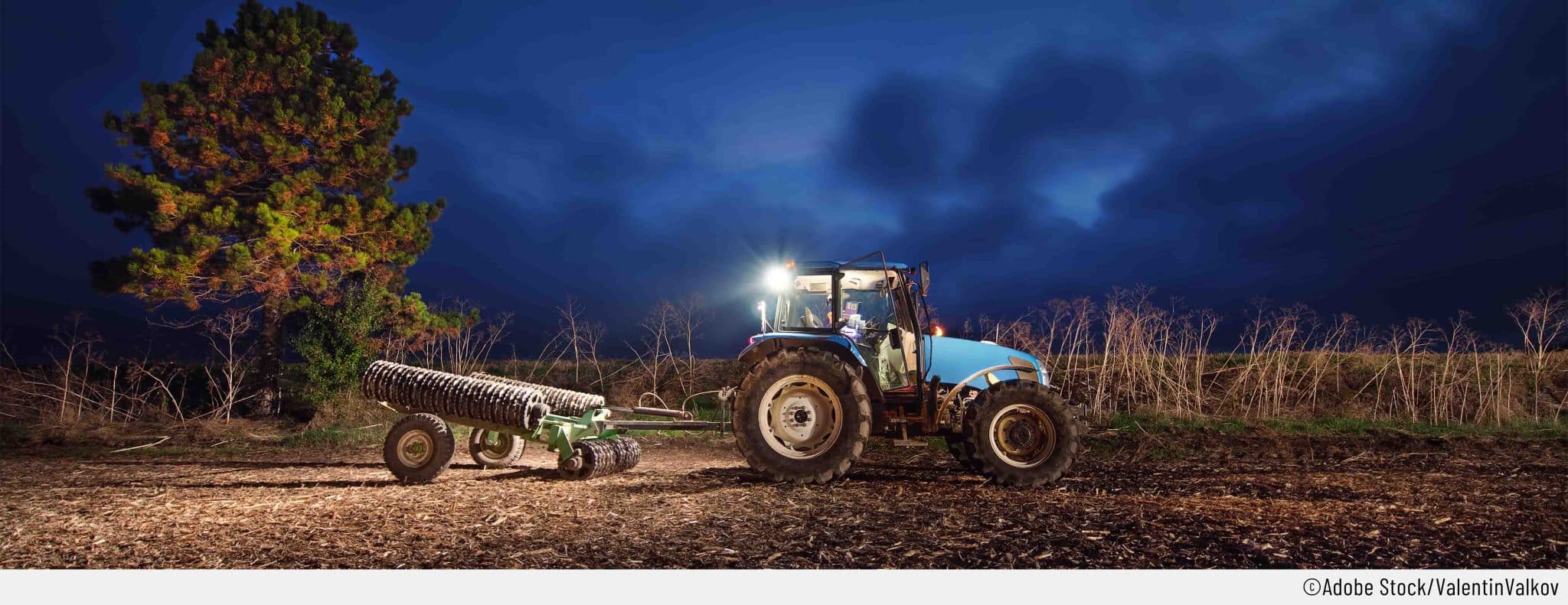 Ein Traktor in der Dämmerung auf dem Feld mit Anhänger und Arbeitsscheinwerfern.