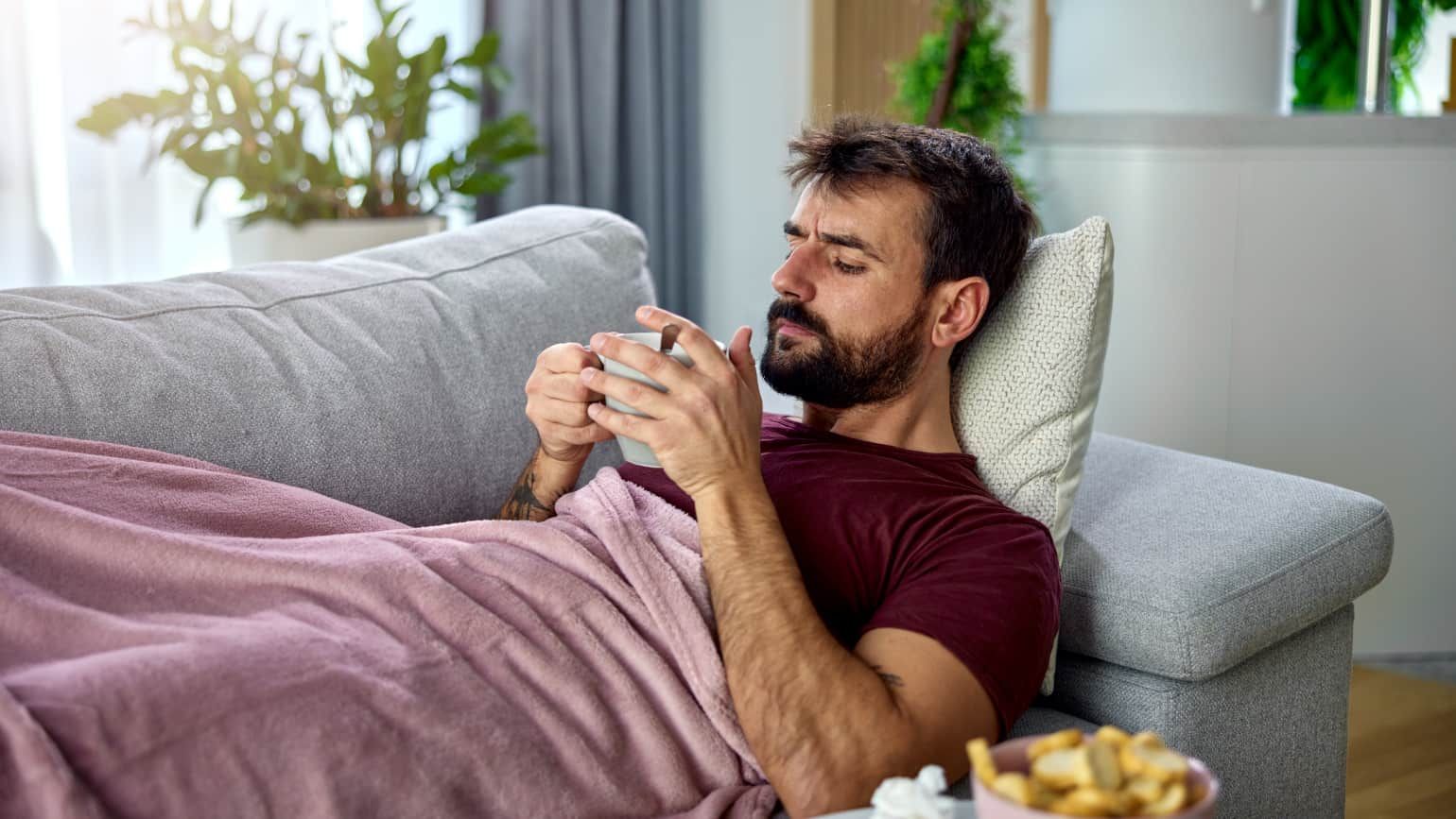 Ein krank wirkender Mann liegt auf einem Sofa. Er ist in eine Decke eingehüllt und hält eine Tasse in der Hand.