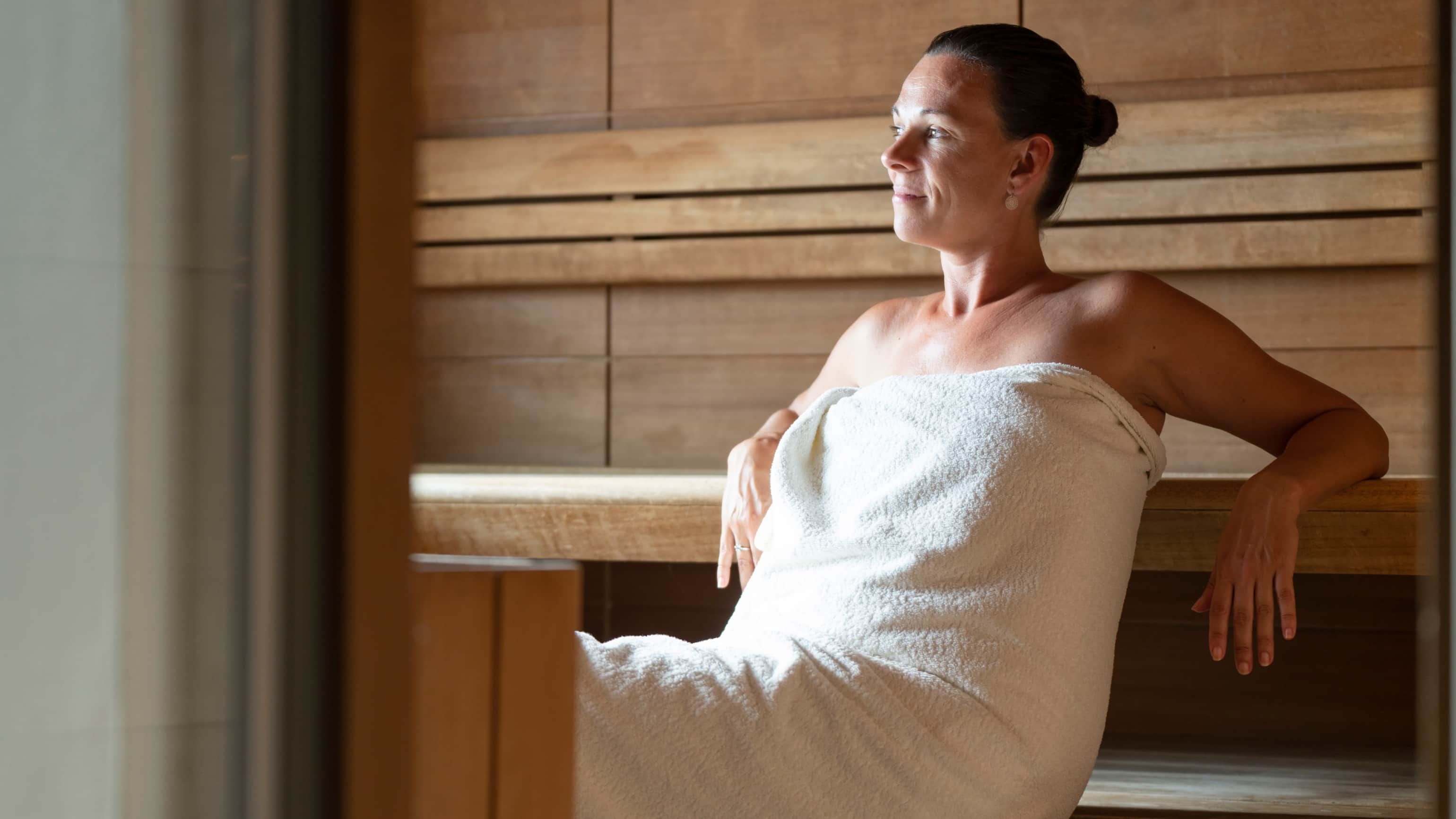 Eine in ein Badetuch gehüllte Frau sitzt entspannt in einer Sauna.