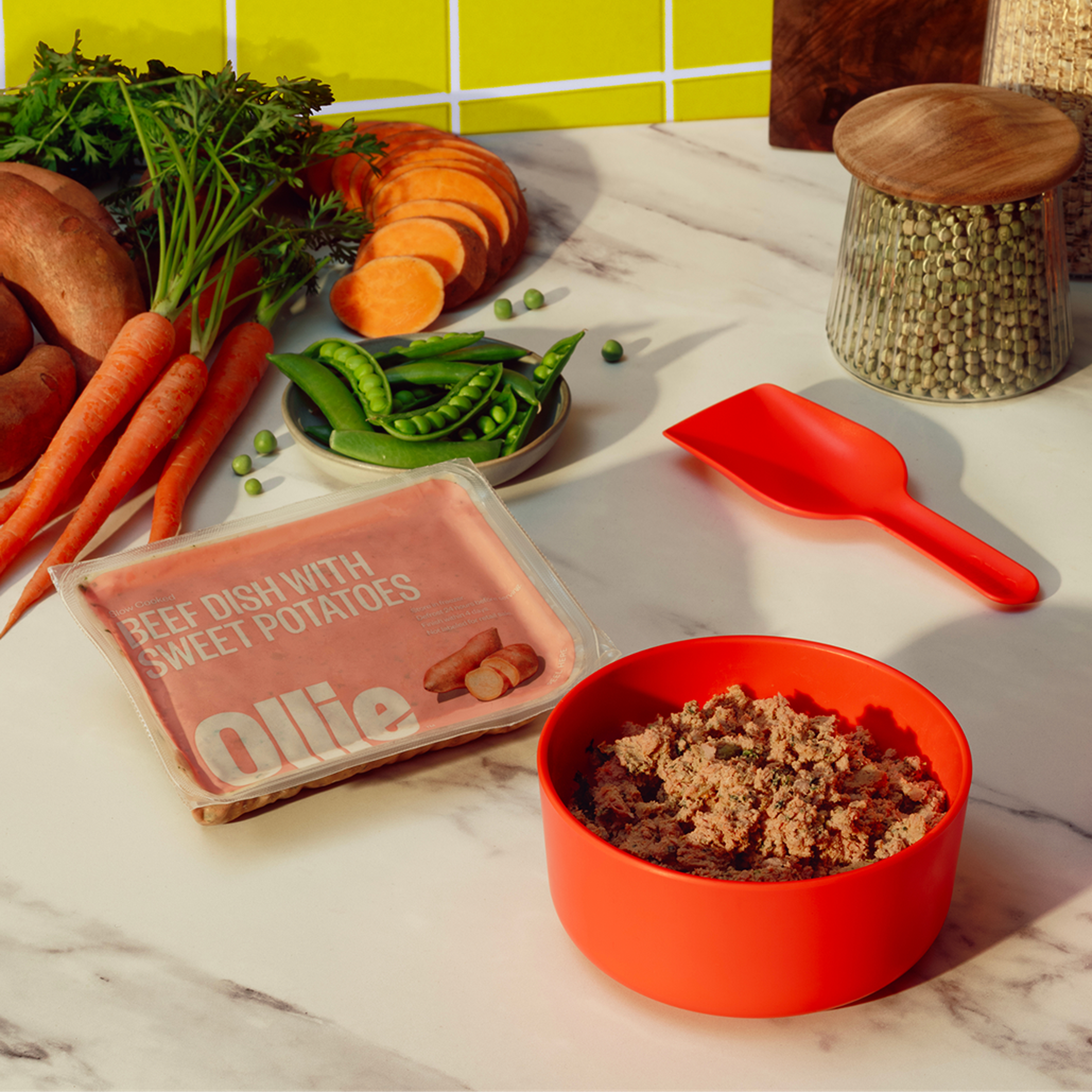 Close-up of a meal prep with a red bowl of Ollie fresh beef dish, sweet potatoes, carrots, peas, and a sealed package labeled "Ollie" on a kitchen counter.