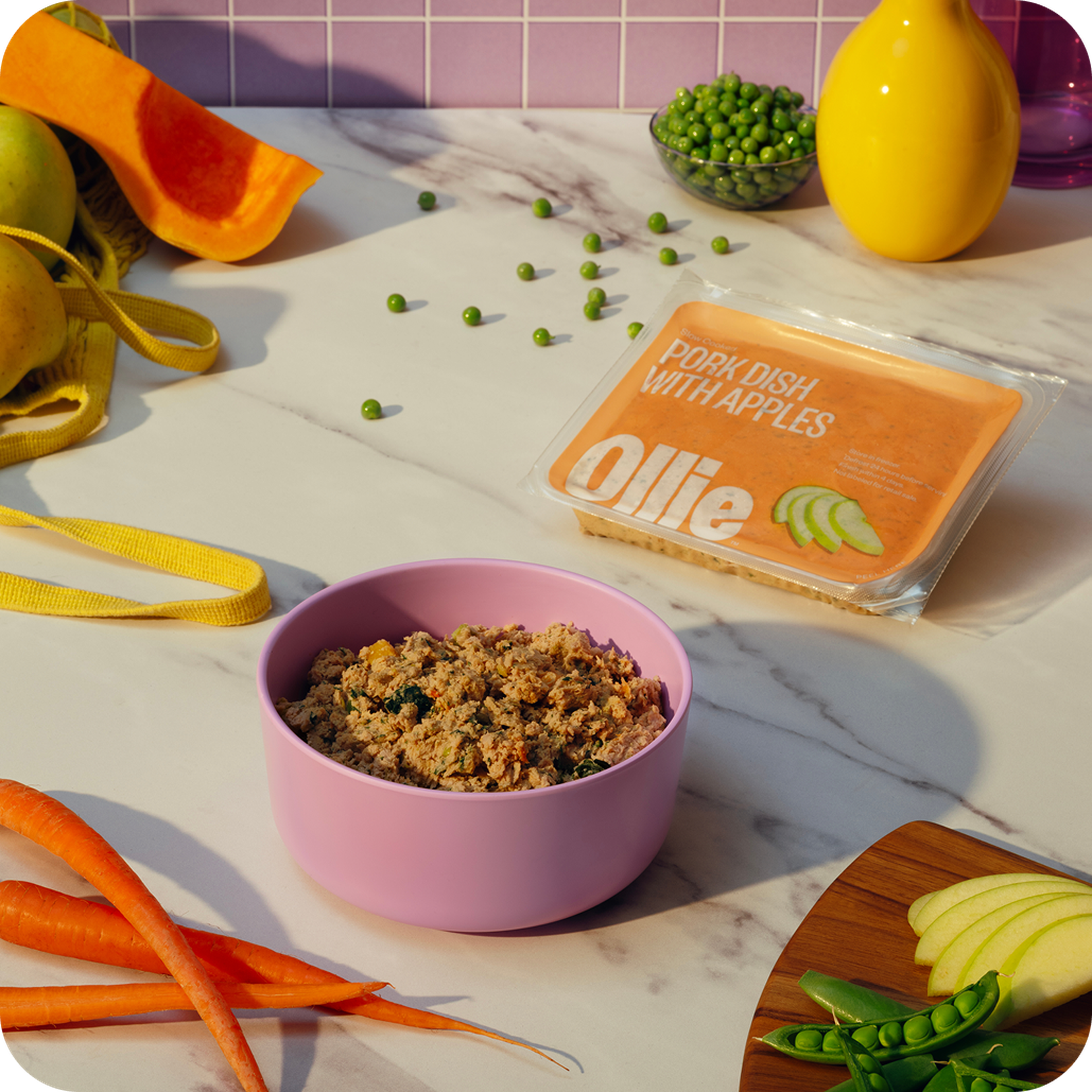 A purple bowl of dog food next to an Ollie package labeled "Pork Dish with Apples" on a marble surface, surrounded by peas, carrots, and apples.