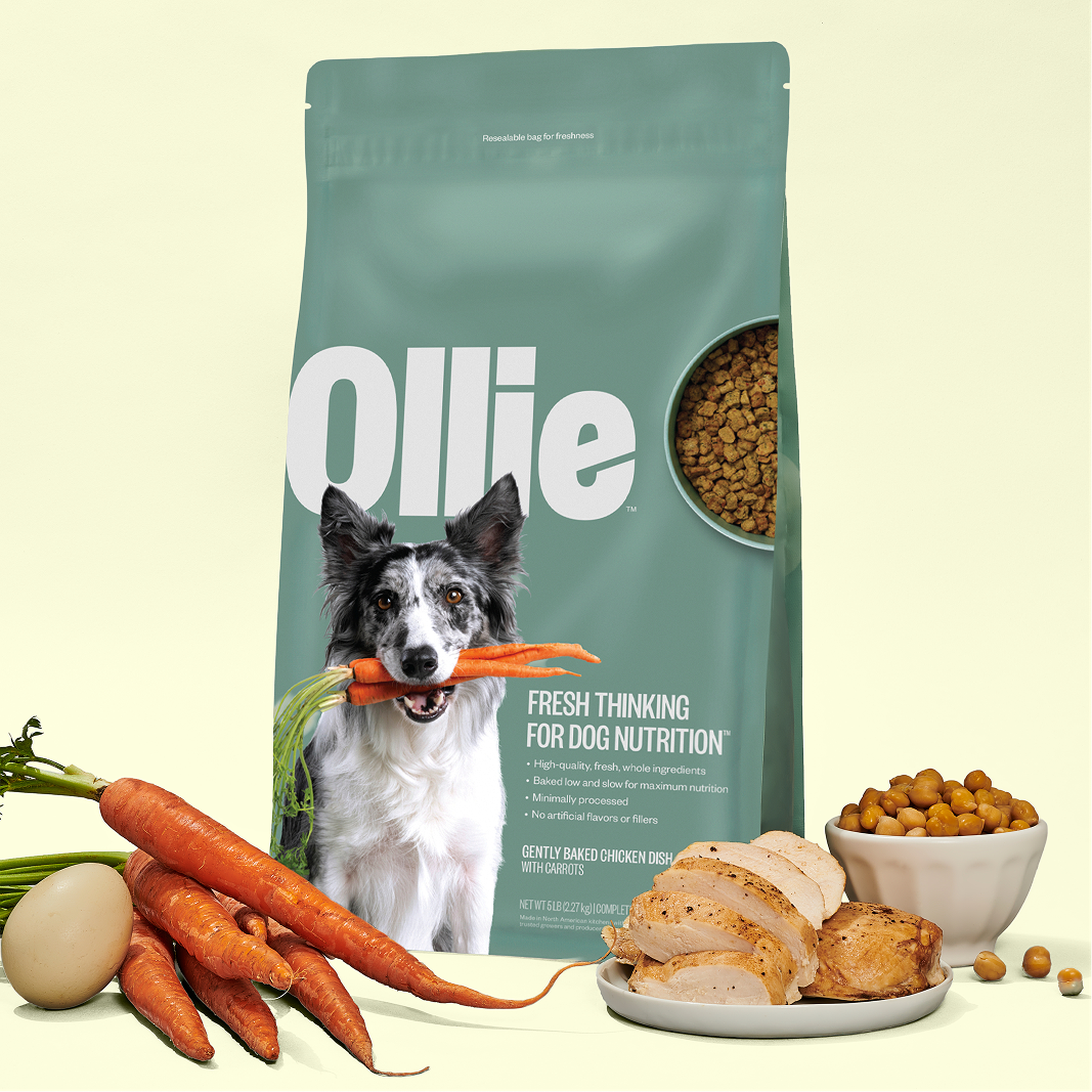 Baked dog food bag labeled "Ollie" with image of a dog holding a carrot. Surrounding ingredients: carrots, egg, chickpeas, and cooked chicken slices.