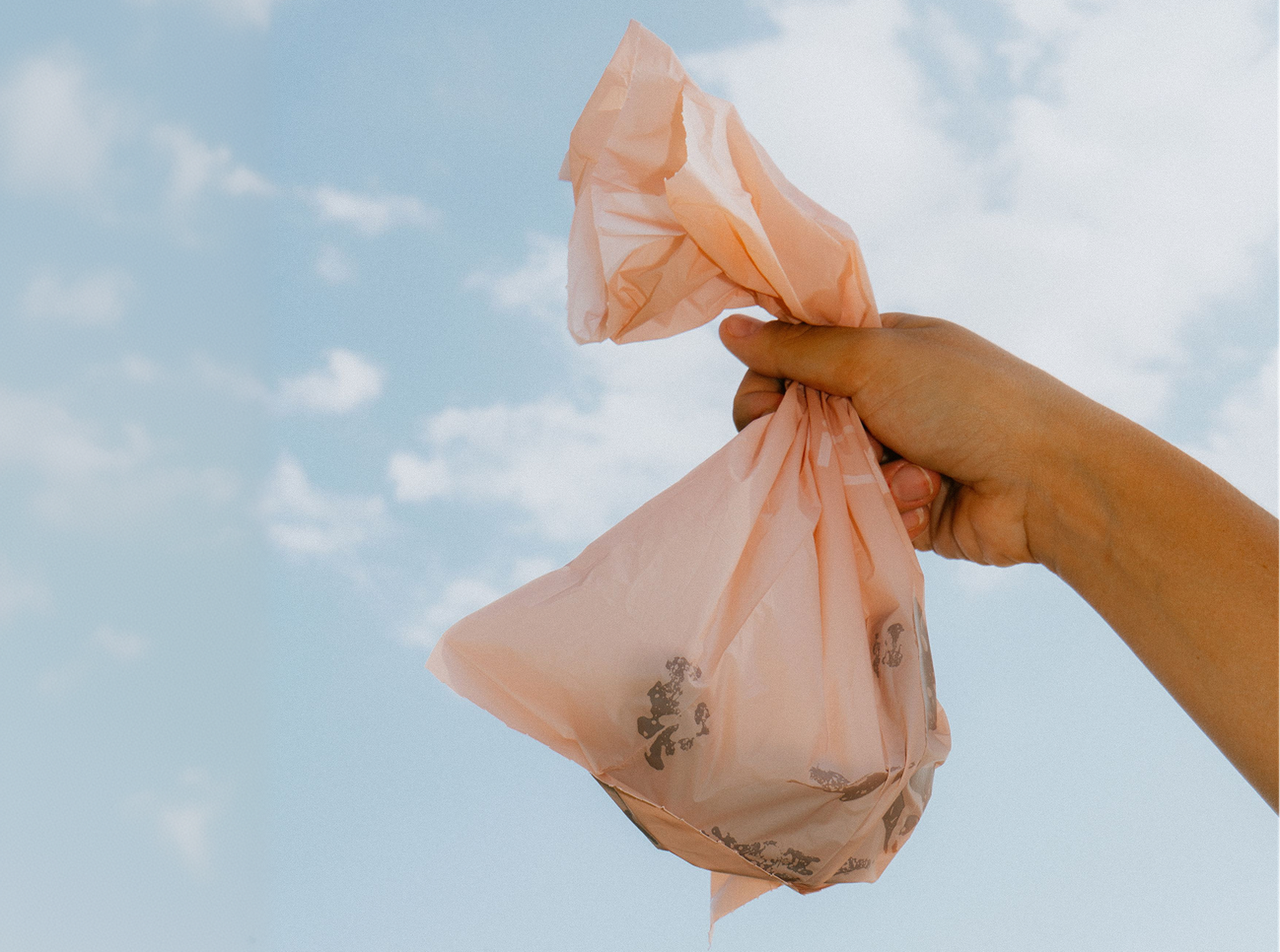 Hand holding a crumpled pink plastic bag against a clear blue sky with scattered clouds.