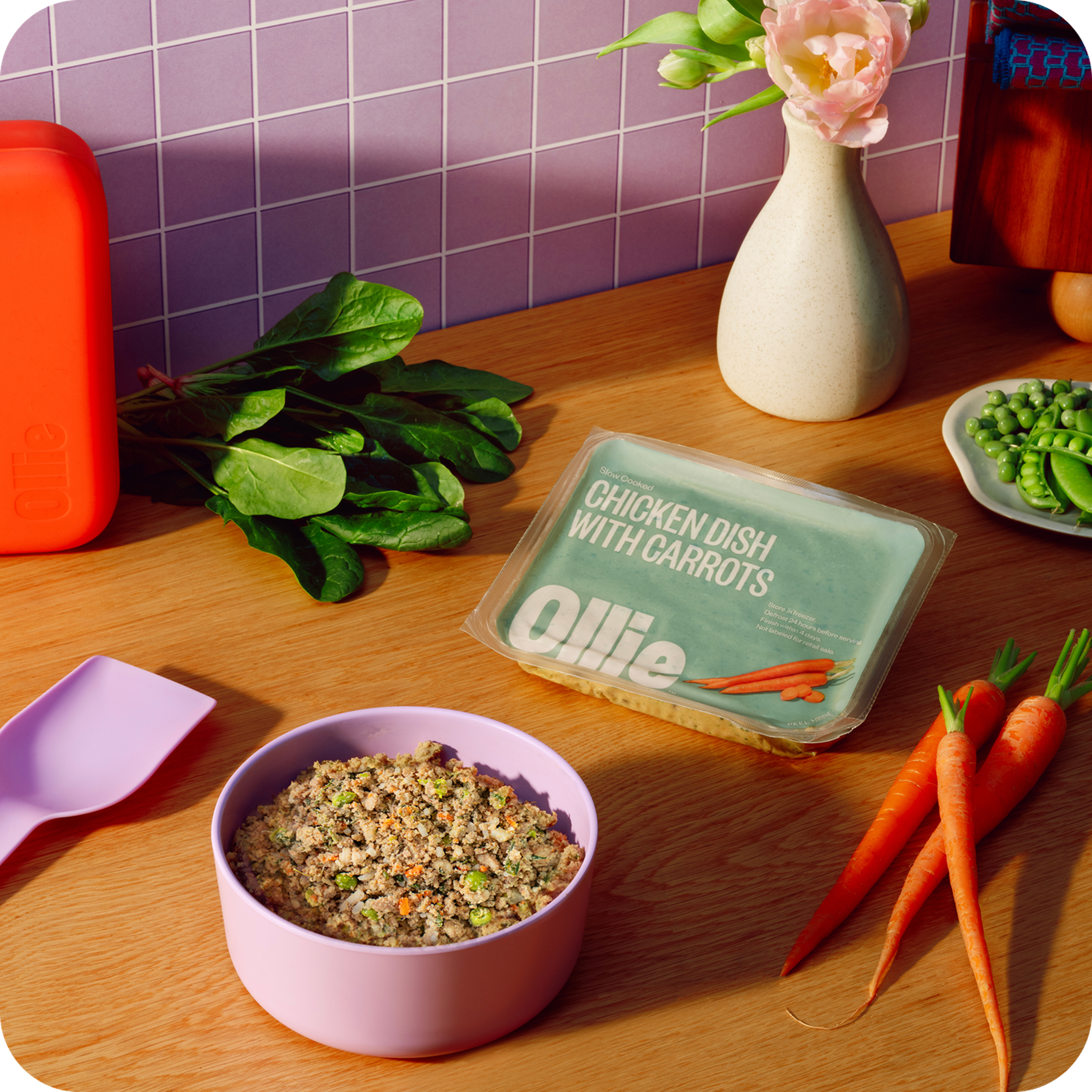 A bowl of chicken and carrot dish in pink bowl, with fresh carrots, spinach, peas, a pink flower in vase, and an orange container nearby on a wooden table.