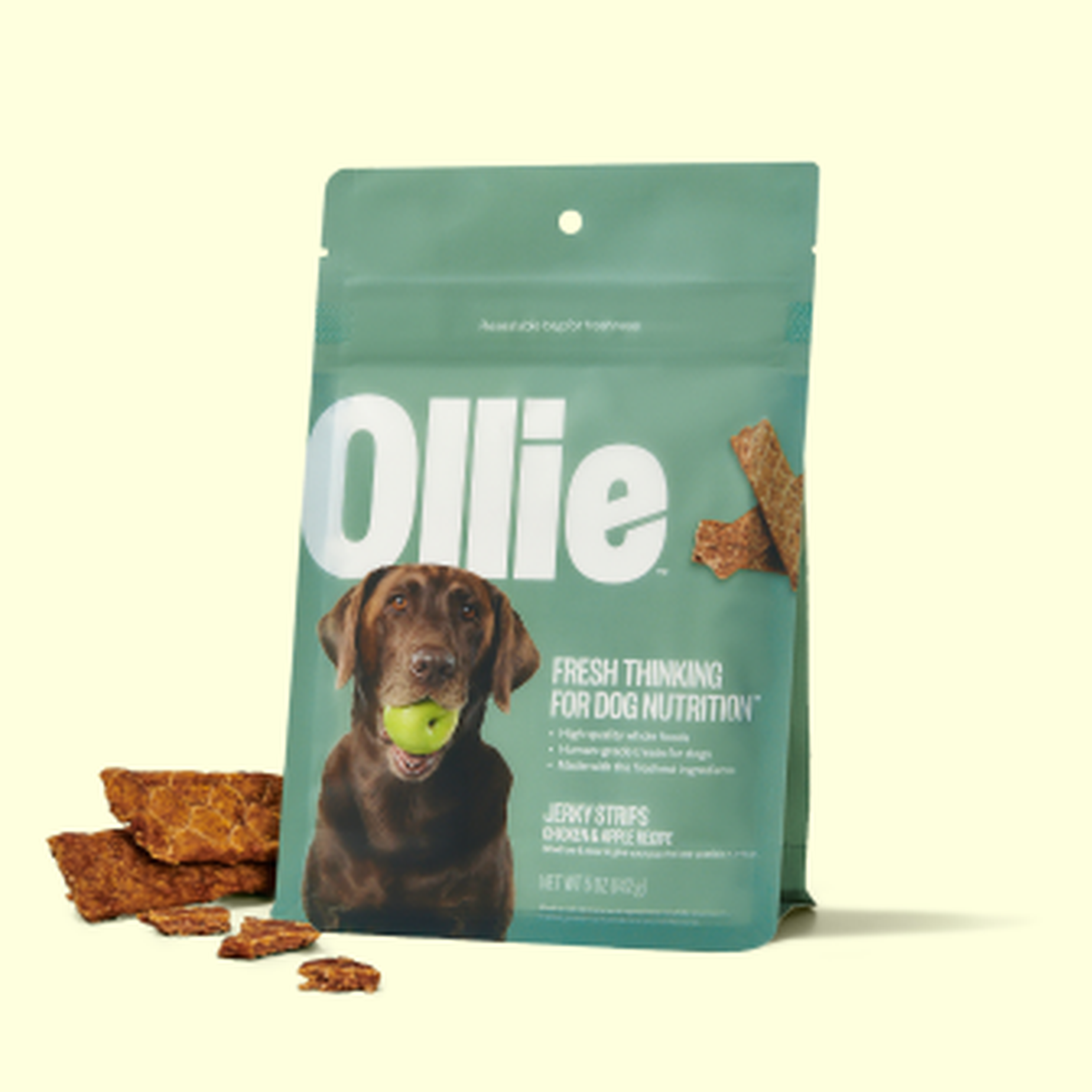Dog treat package labeled "Ollie" with a picture of a dog holding an apple. Jerky strips scattered nearby.