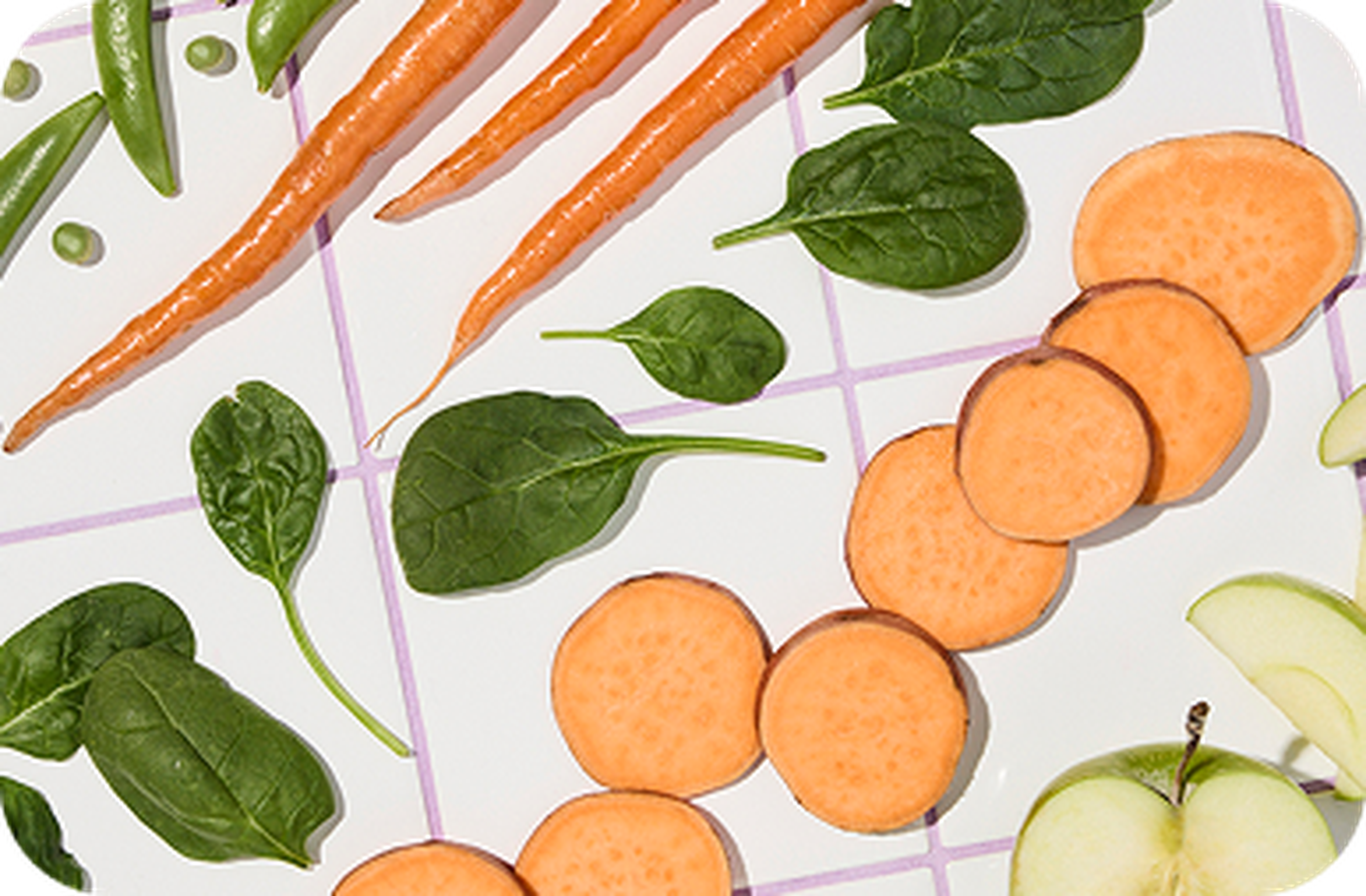 Fresh vegetables and fruits arranged on a patterned surface, including carrots, spinach leaves, sweet potato slices, and apple pieces.