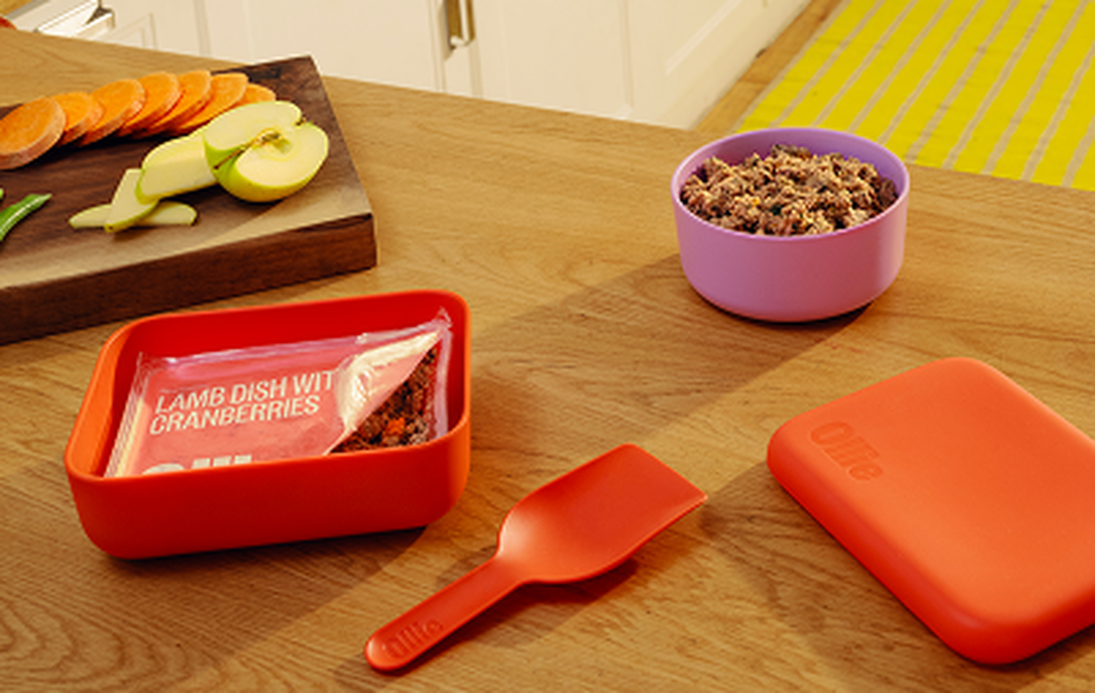 Red container of lamb with cranberries, bowl of Ollie fresh dog food, and a scoop on a wooden counter with sliced vegetables nearby.