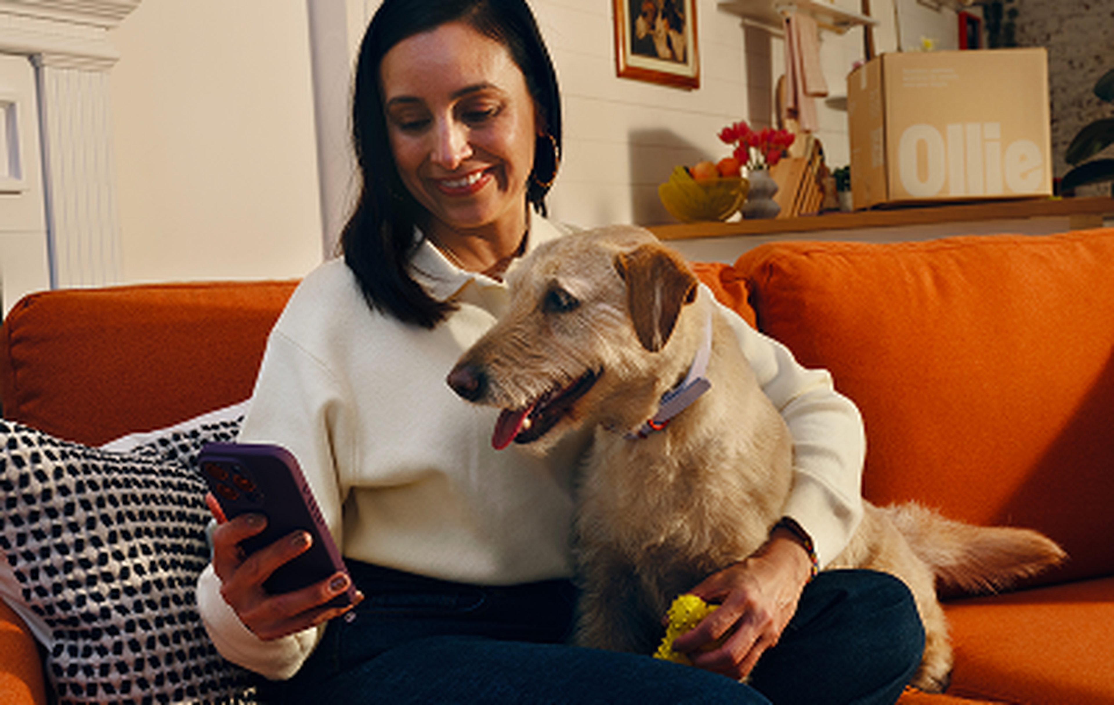 A woman sits on an orange sofa, smiling and looking at her phone, with a dog cuddling beside her, holding a yellow toy.