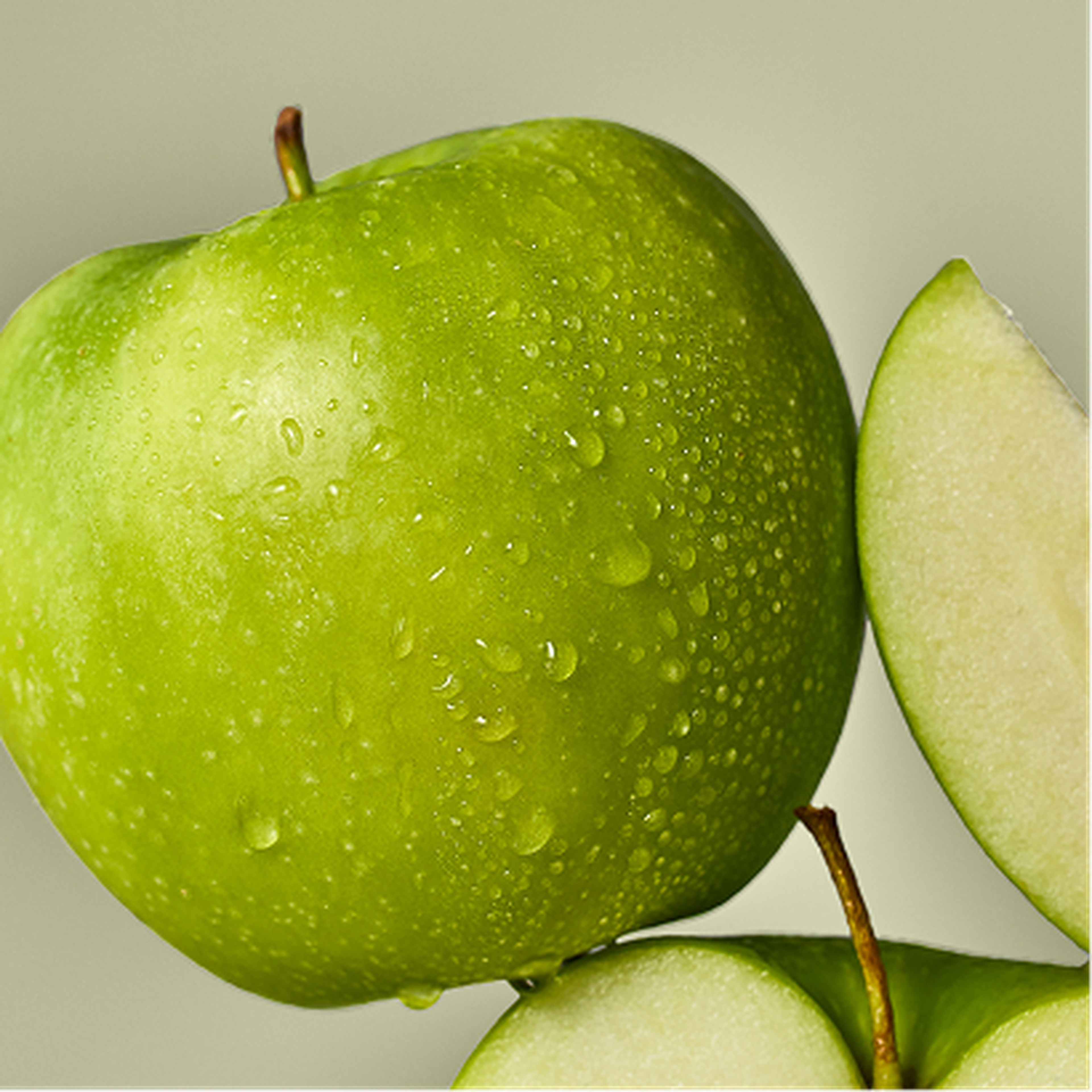Close-up of a fresh green apple with water droplets, next to a sliced piece, against a light green background.