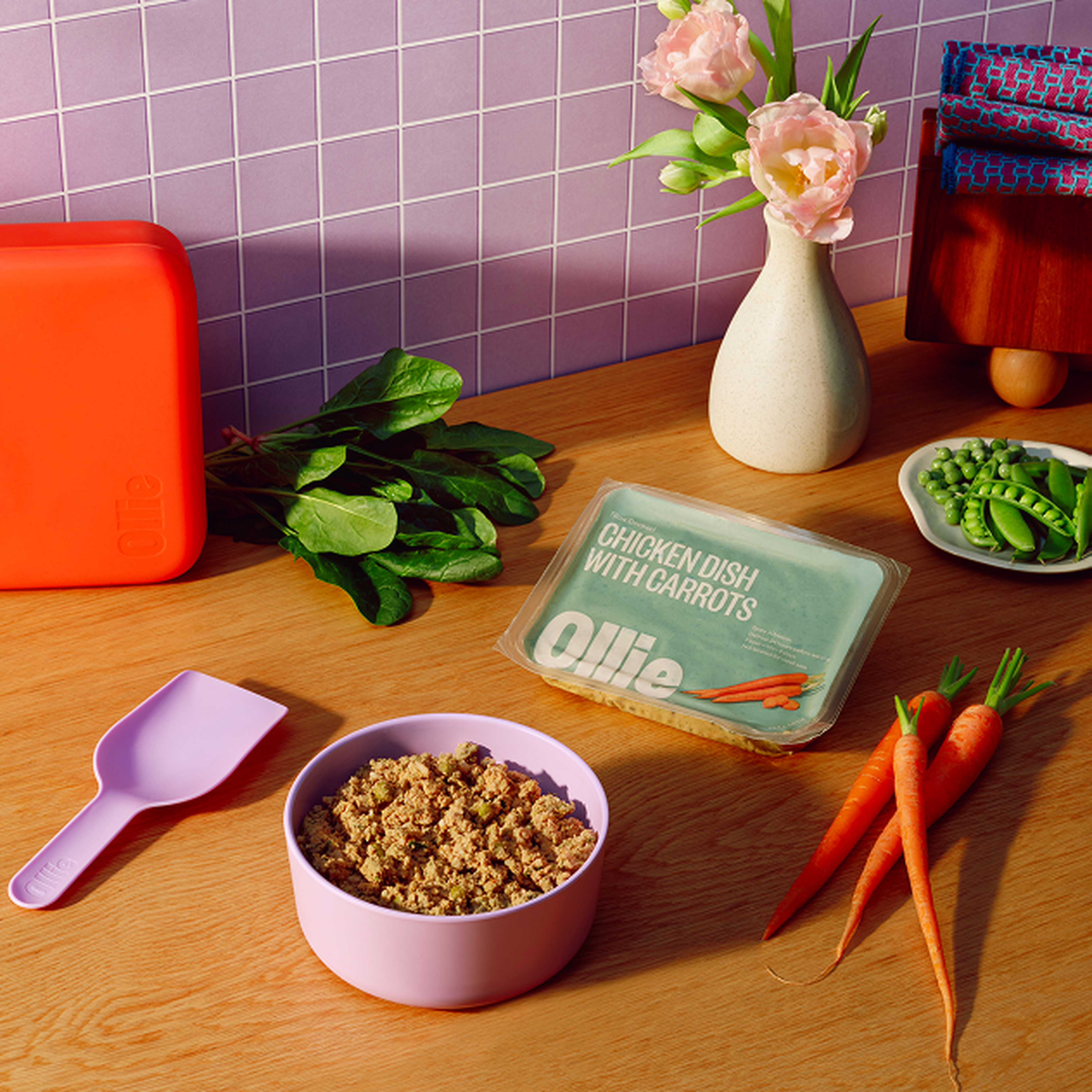 A kitchen scene with a bowl of food, a plastic container labeled "Chicken Dish with Carrots," carrots, a scoop, and a vase with pink flowers.
