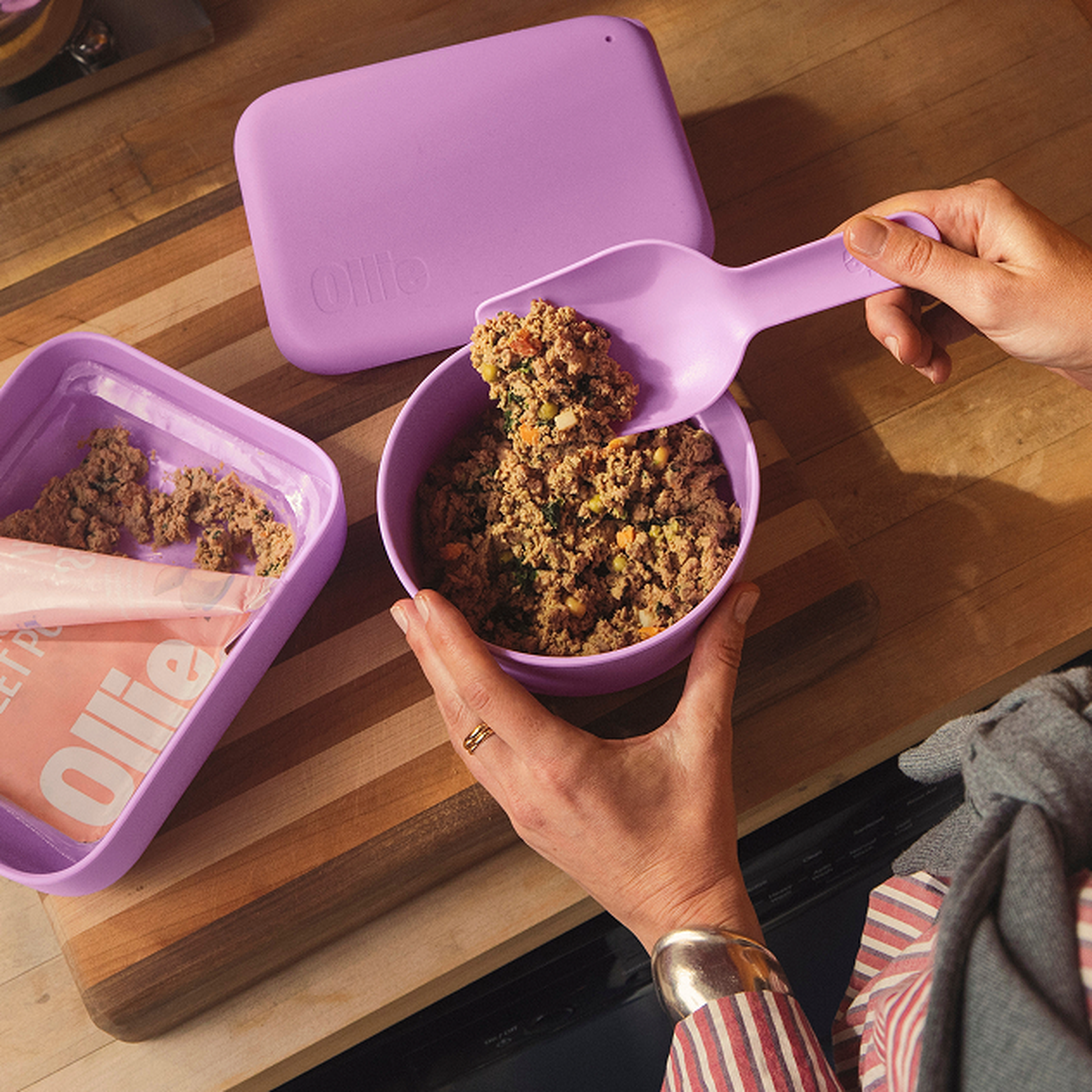 A person scoops dog food from a purple container into a purple bowl on a wooden countertop, with a branded dog food pouch nearby.