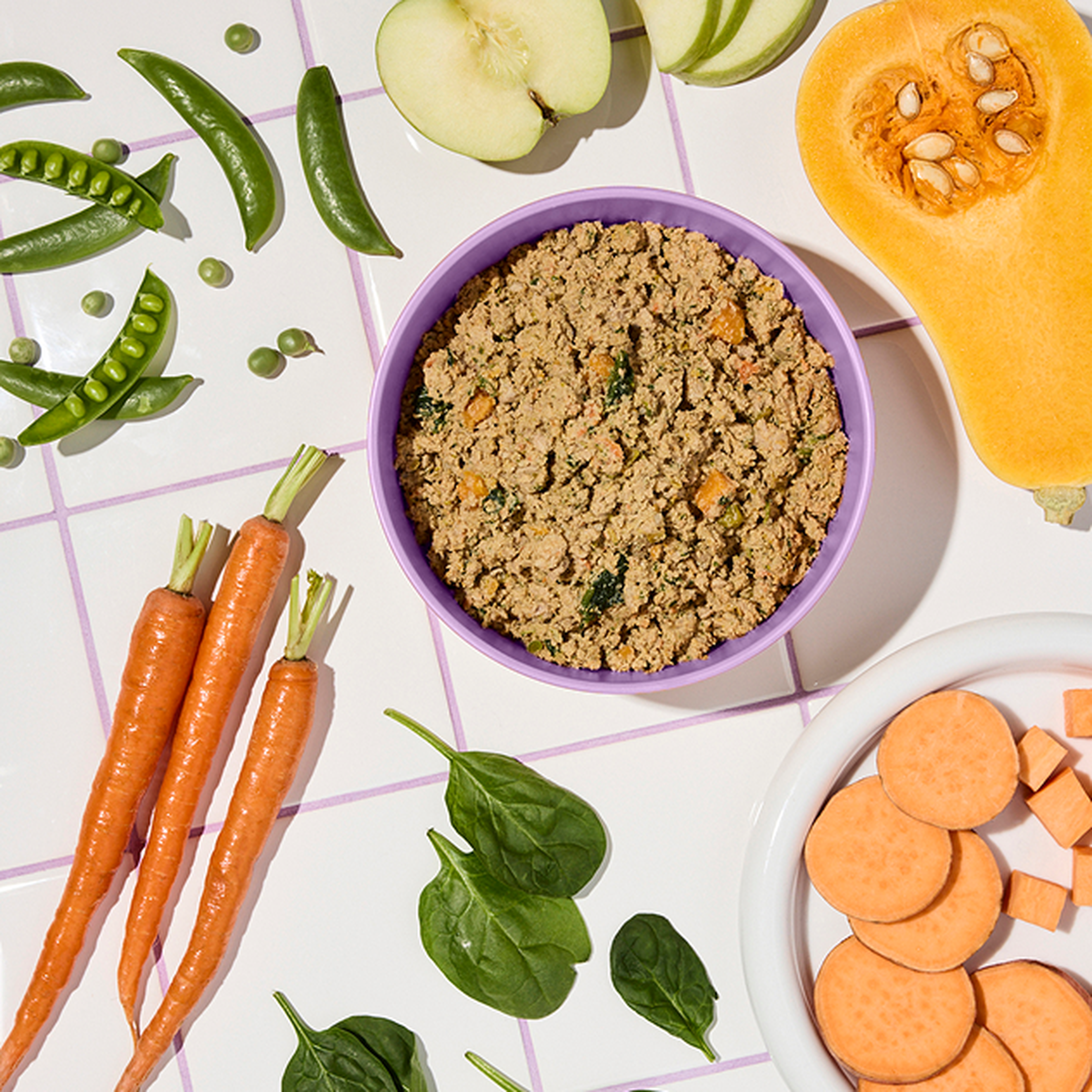 A bowl of fresh Ollie surrounded by carrots, spinach, sliced apple, snap peas, butternut squash, and sweet potatoes on a tiled surface.