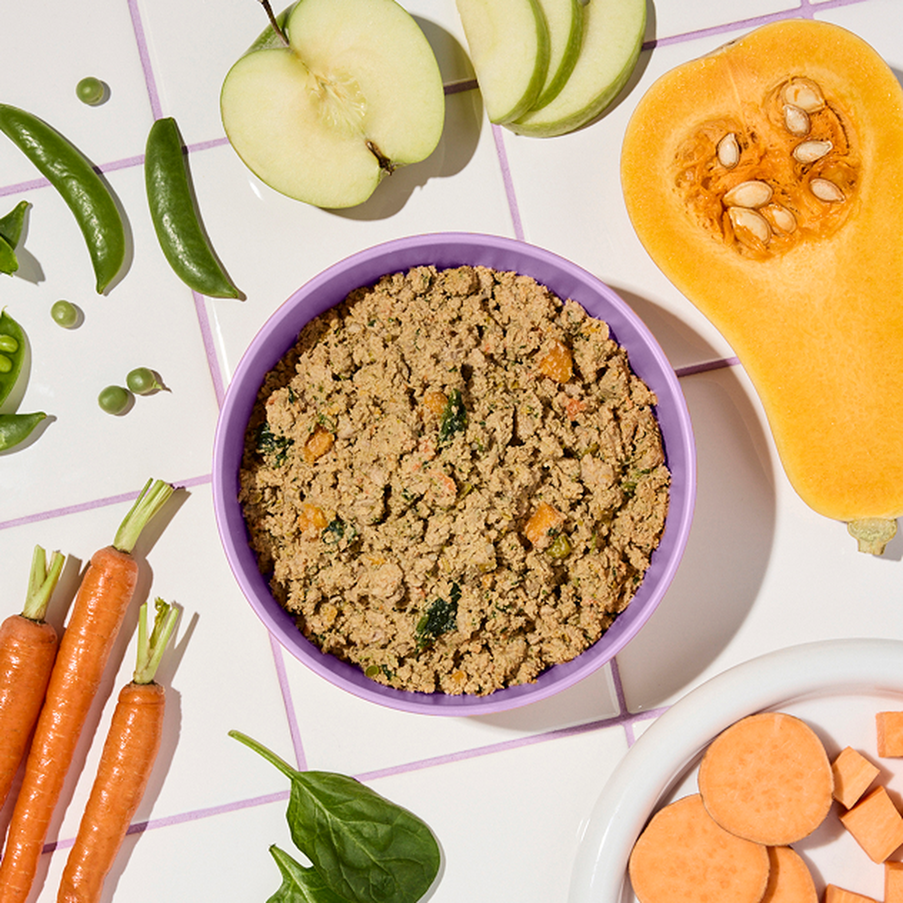 A purple bowl of fresh Ollie, surrounded by fresh ingredients like apple slices, peas, spinach, carrots, and squash on a tiled surface.