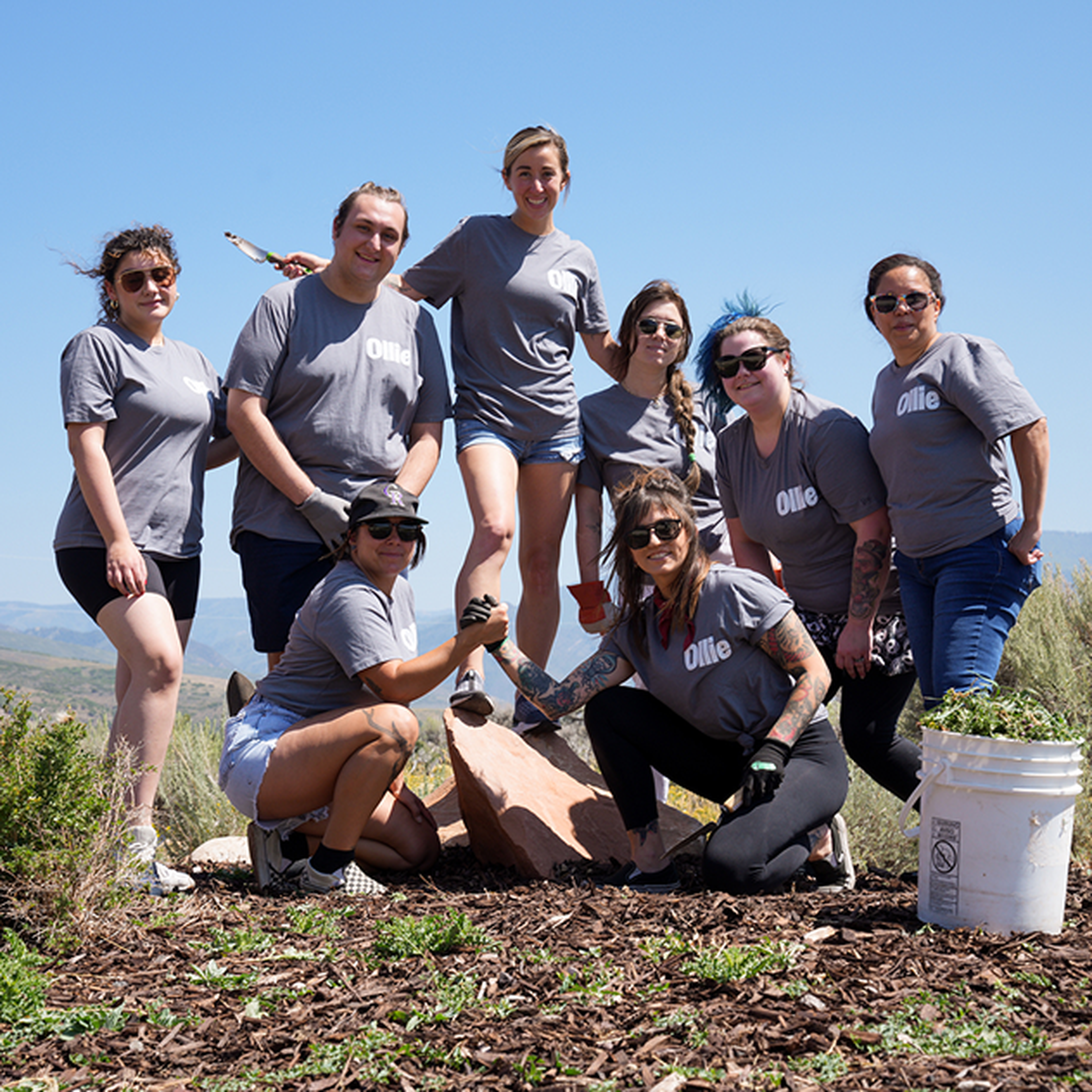 A group of eight people in matching T-shirts posing outdoors on a sunny day, with gardening tools and a bucket of weeds, smiling at the camera.