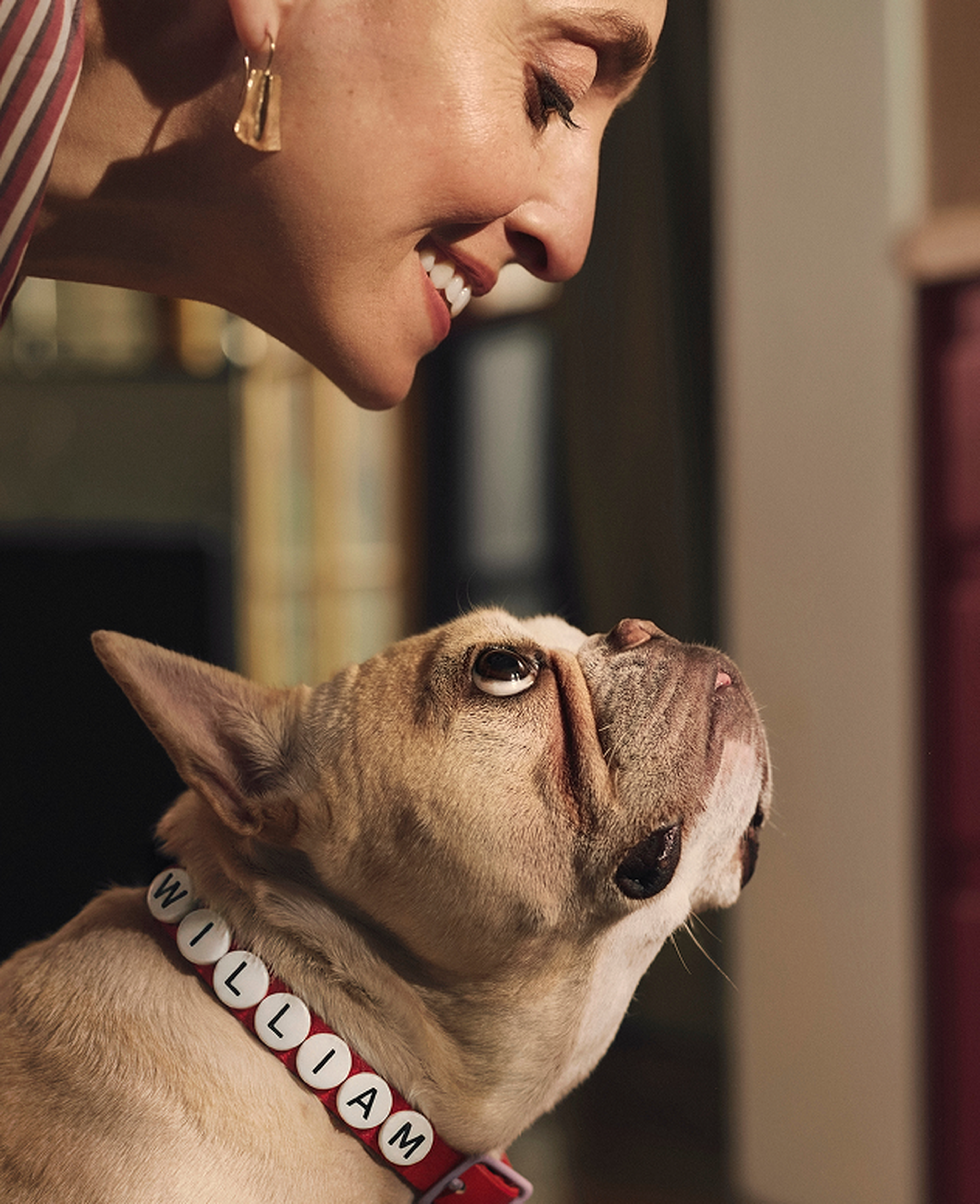 A smiling woman leans down towards a French bulldog wearing a collar with the name "William."