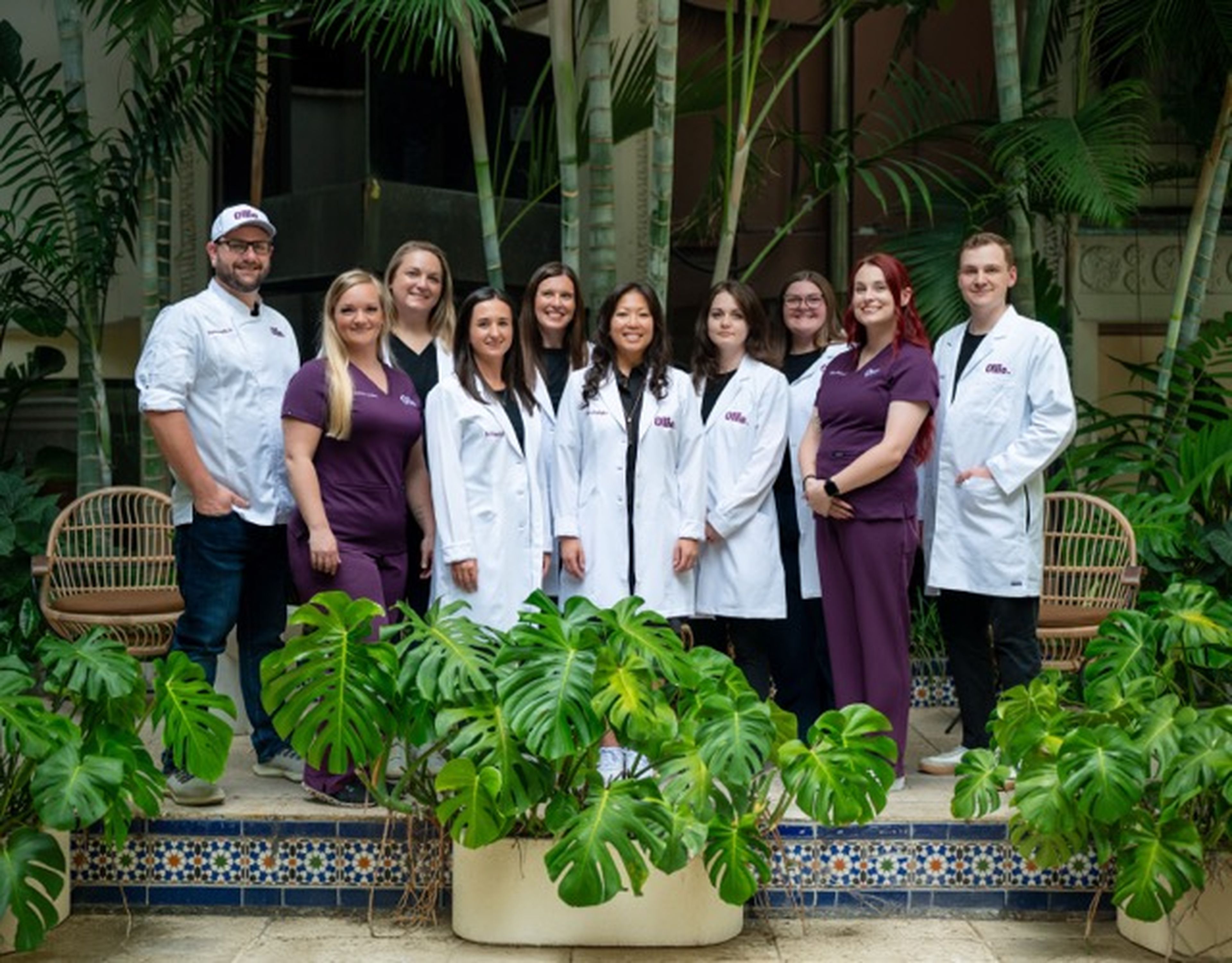 A group of nine people, some in white coats and others in purple scrubs, pose together indoors surrounded by lush green plants.