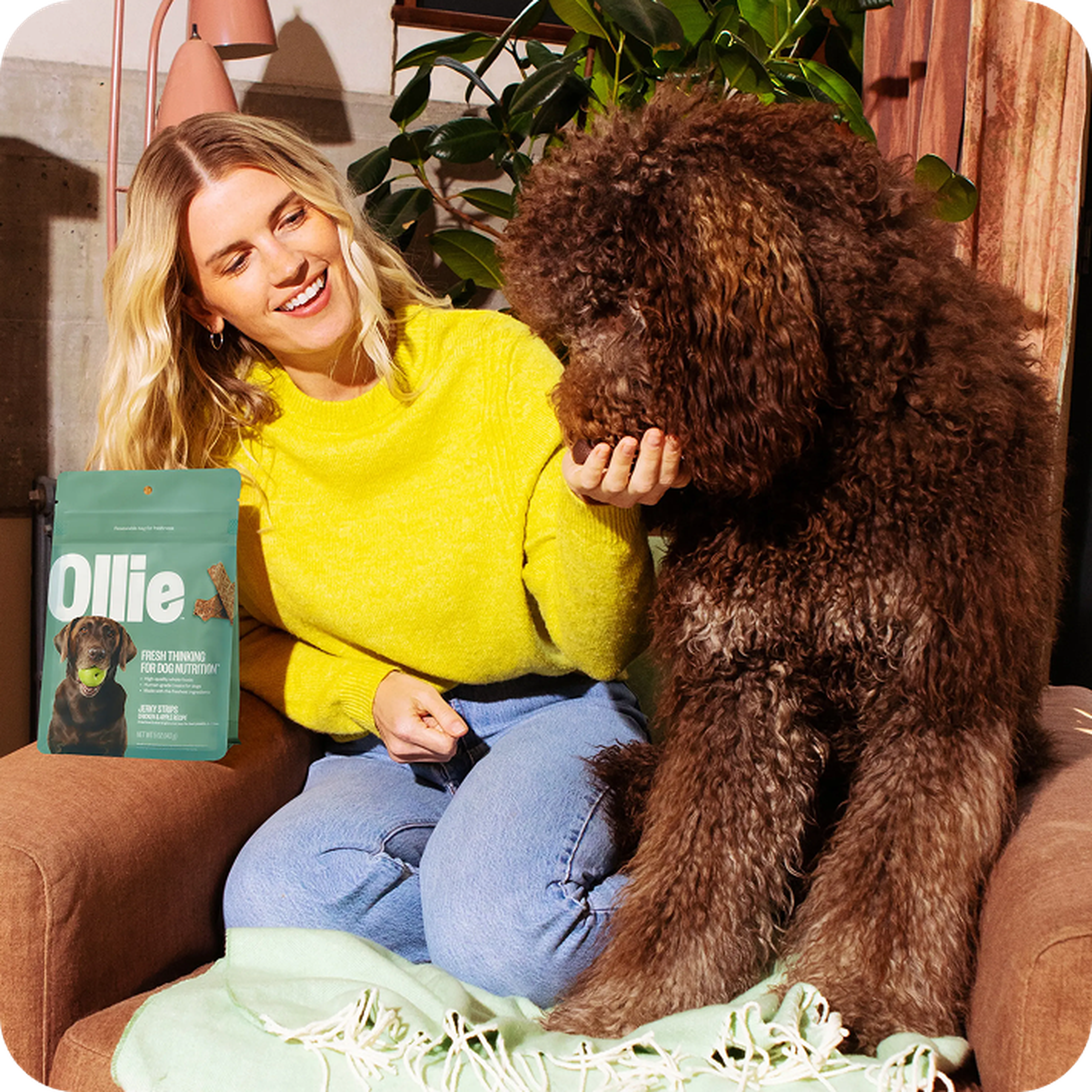 A woman in a yellow sweater sits next to a fluffy brown dog on a couch, holding a dog treat. A pack of dog food is on the side.
