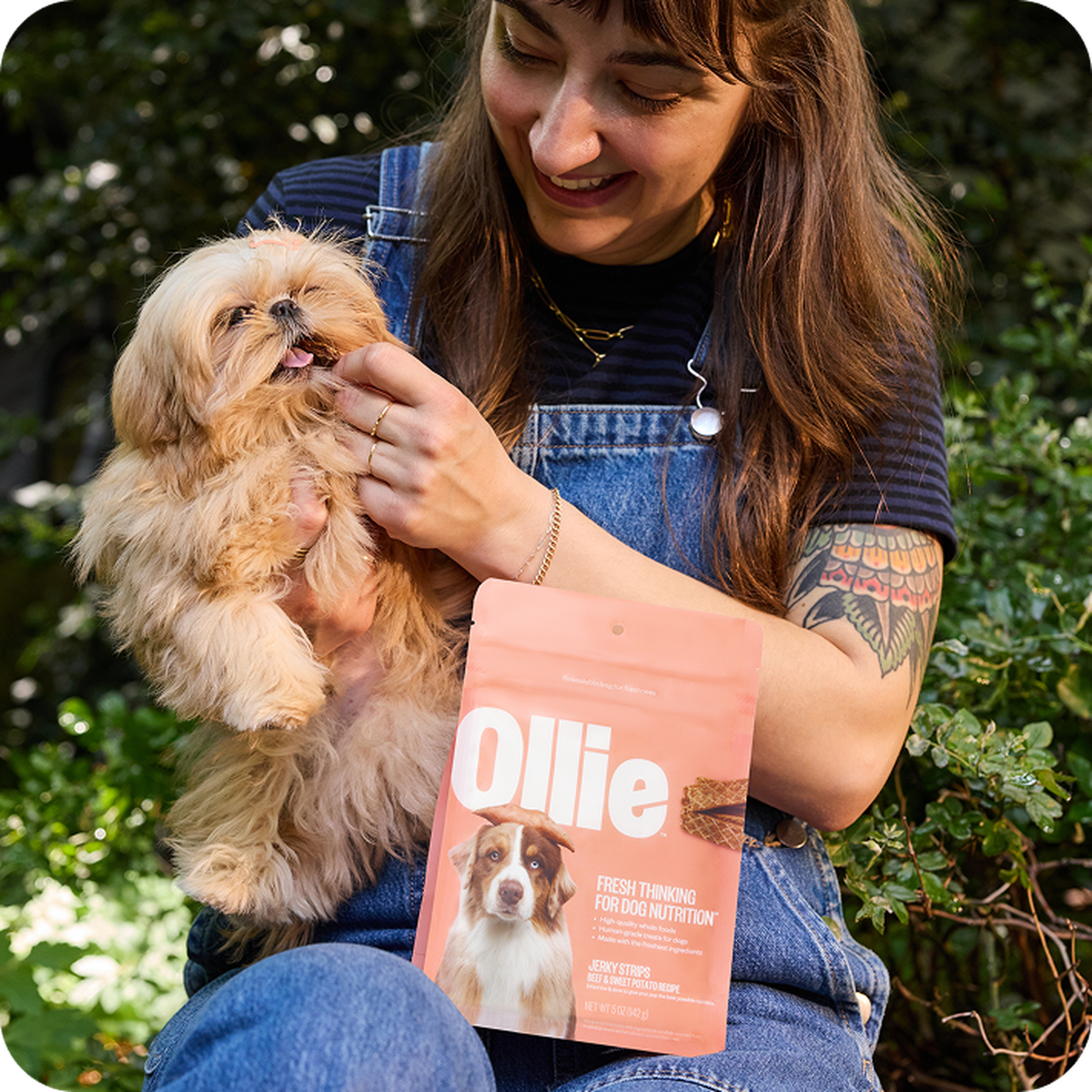 A woman smiles at a small fluffy dog she's holding, while sitting outdoors with a package of Ollie dog food in her lap.