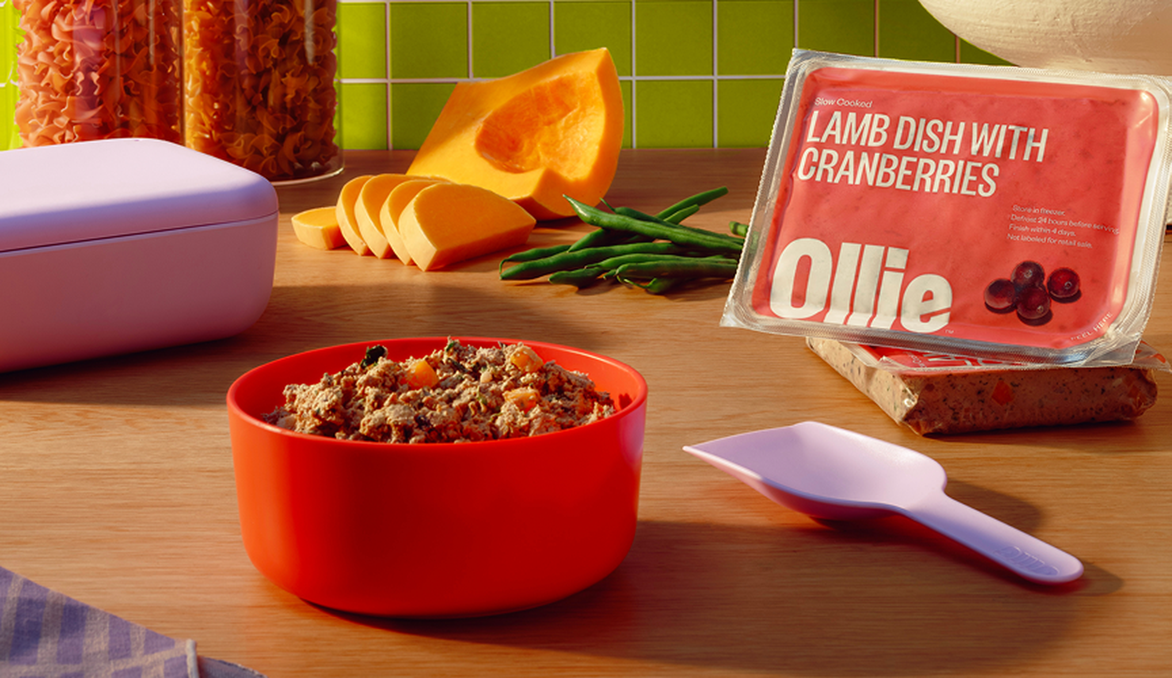 Red bowl of lamb and cranberries next to Ollie food pack, spoon, squash slices, green beans, and pasta on a wooden table.