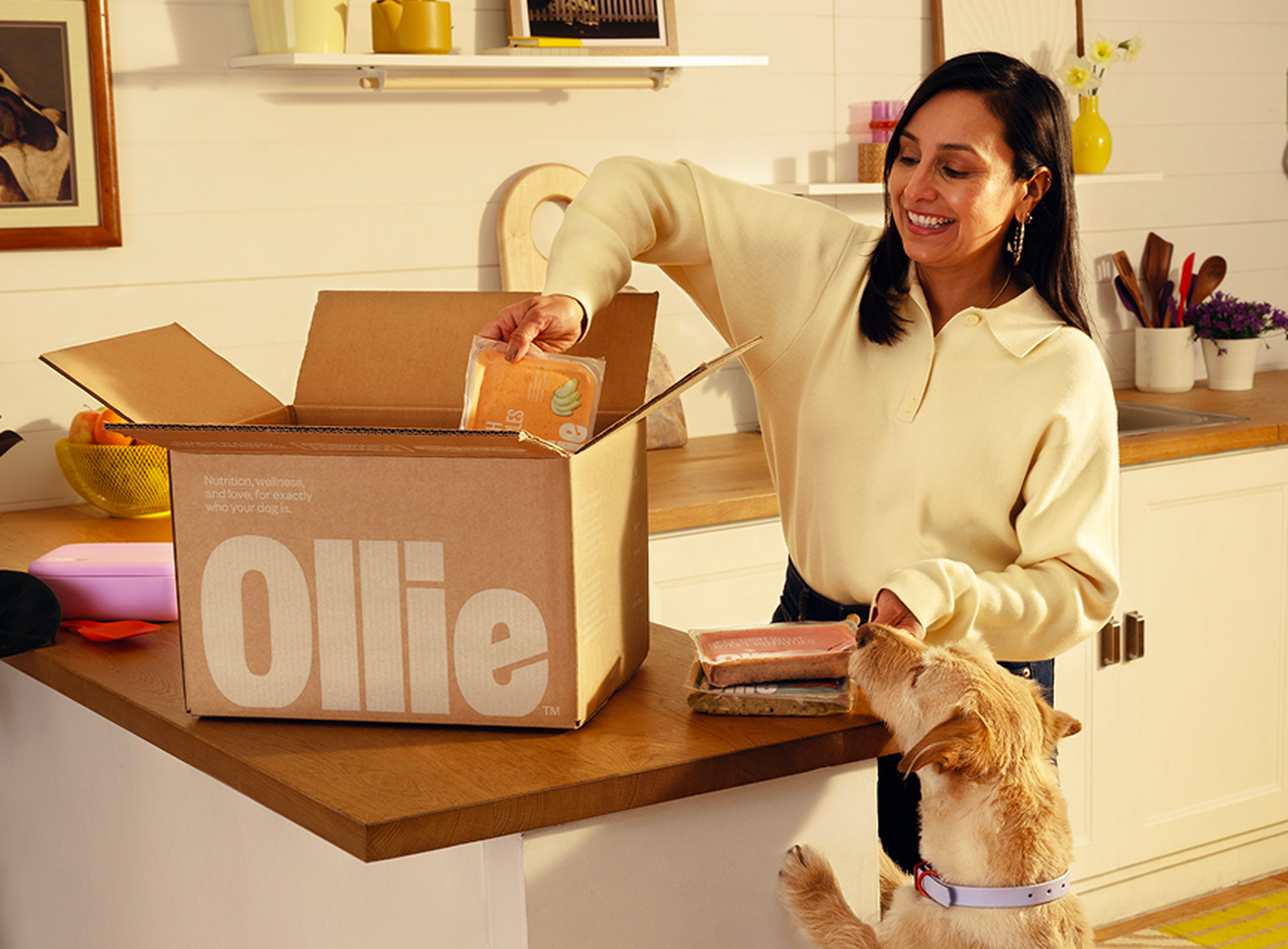 A woman smiles while unpacking a box labeled "Ollie" in a kitchen, with a dog eagerly sniffing a pack of food on the counter.