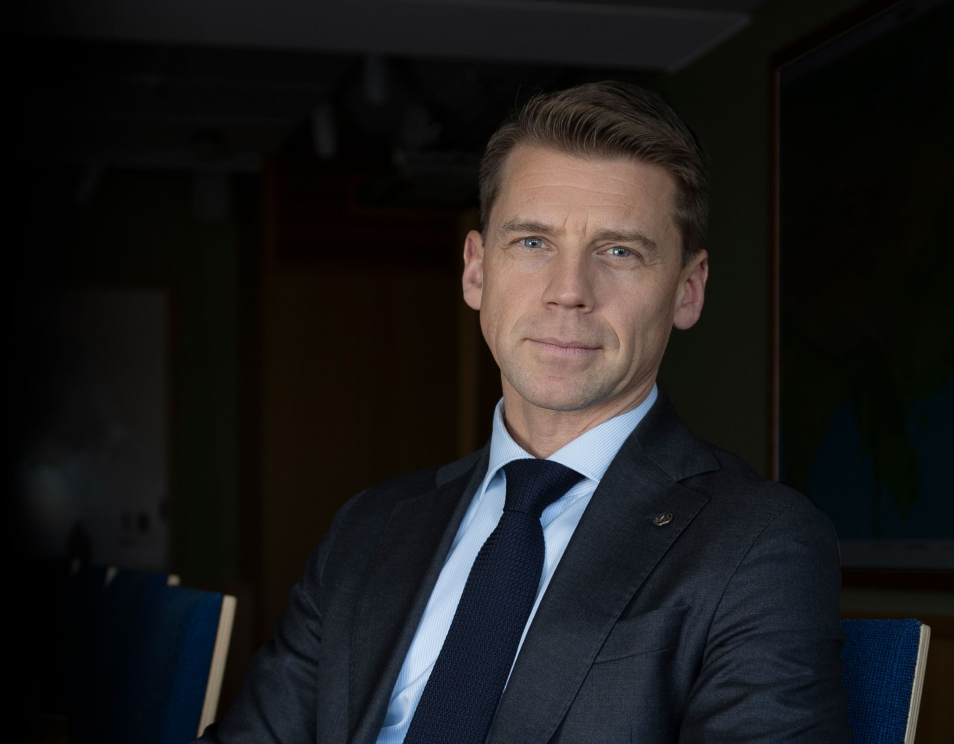 A man in a dark suit and blue tie sits confidently on a chair, hands clasped, with a composed expression in a dimly lit room.