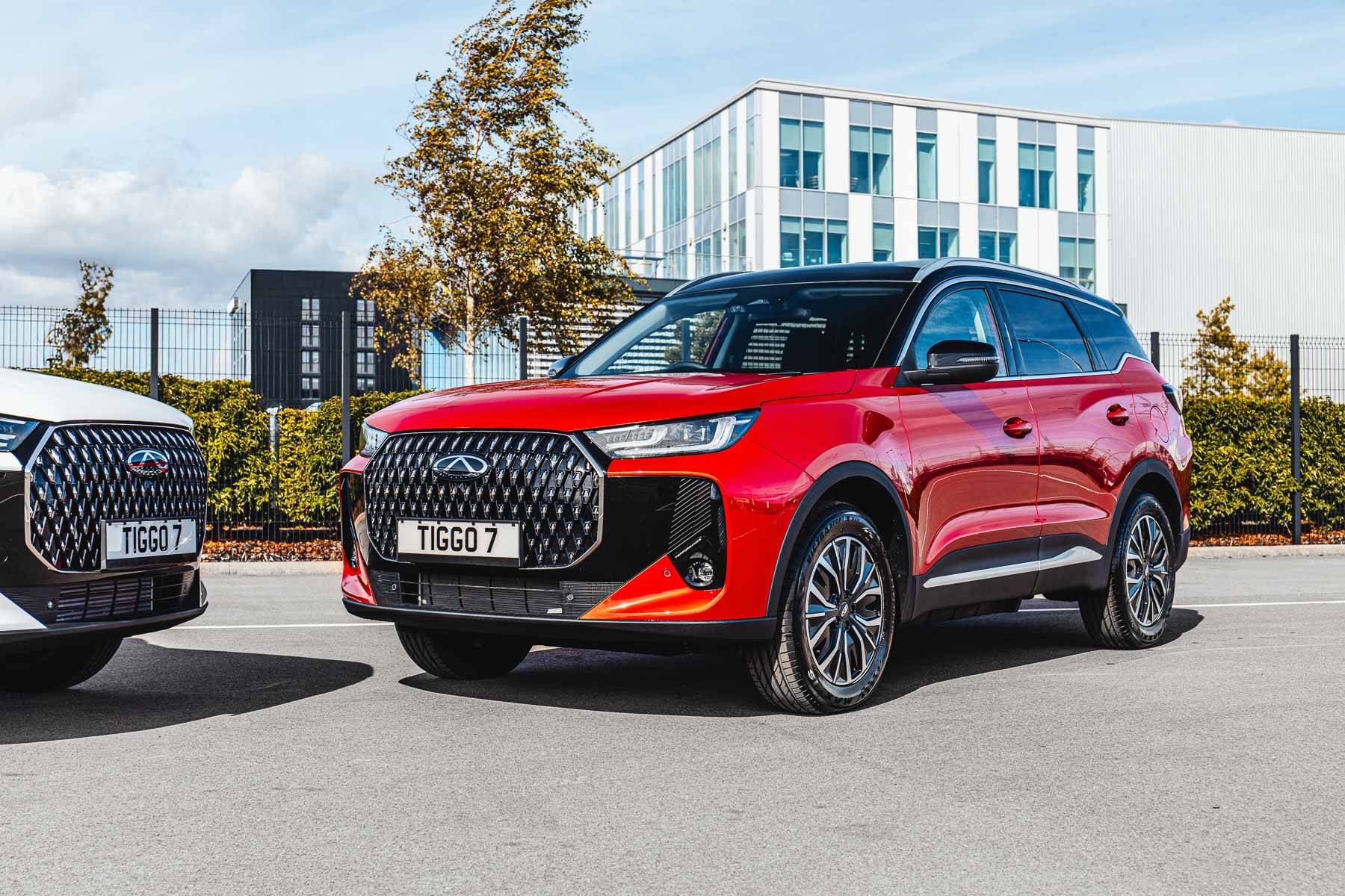 Red Chery TIGGO 7 Hybrid, parked on a sunny day, next to a white vehicle, with modern buildings and trees in the background.