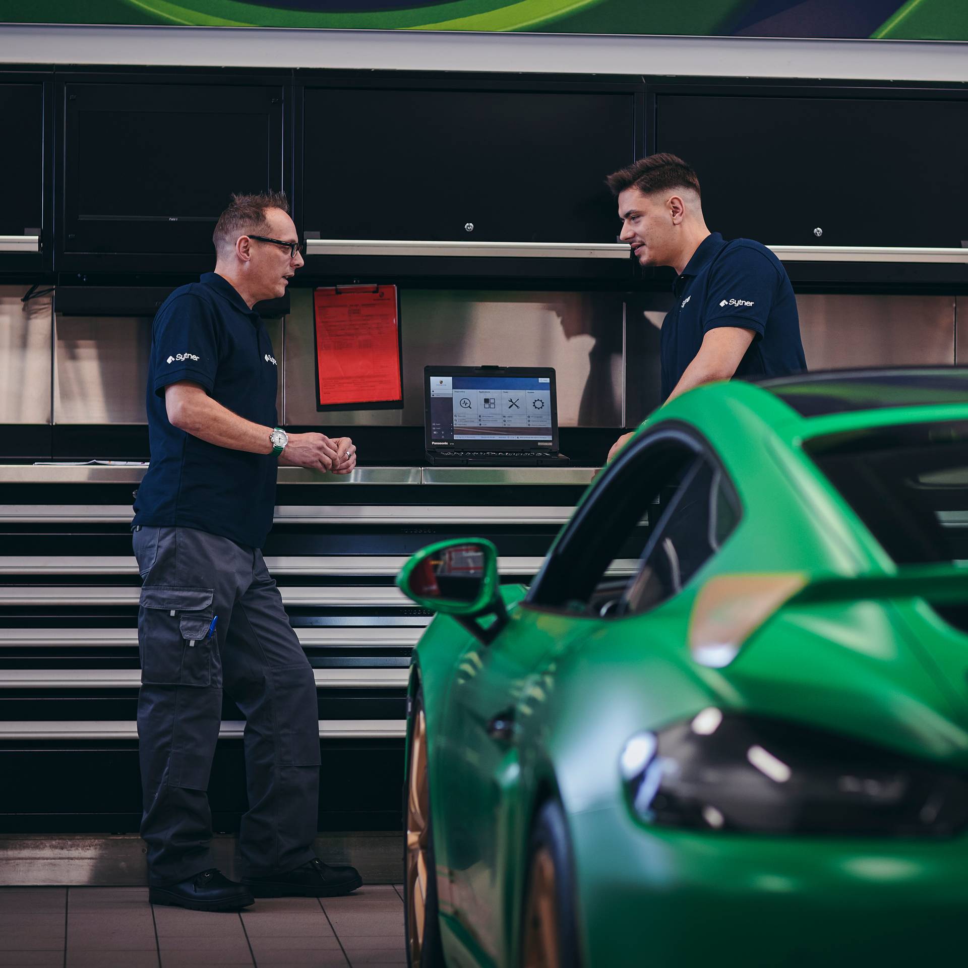 Two technicians talking behind a green Porsche in the workshop