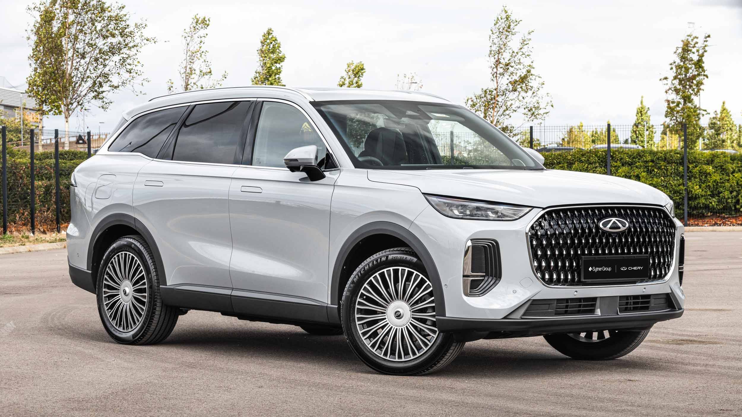 Chery TIGGO 9 front three quarter finished in a silver colour parked in a fenced area with green hedges around it.