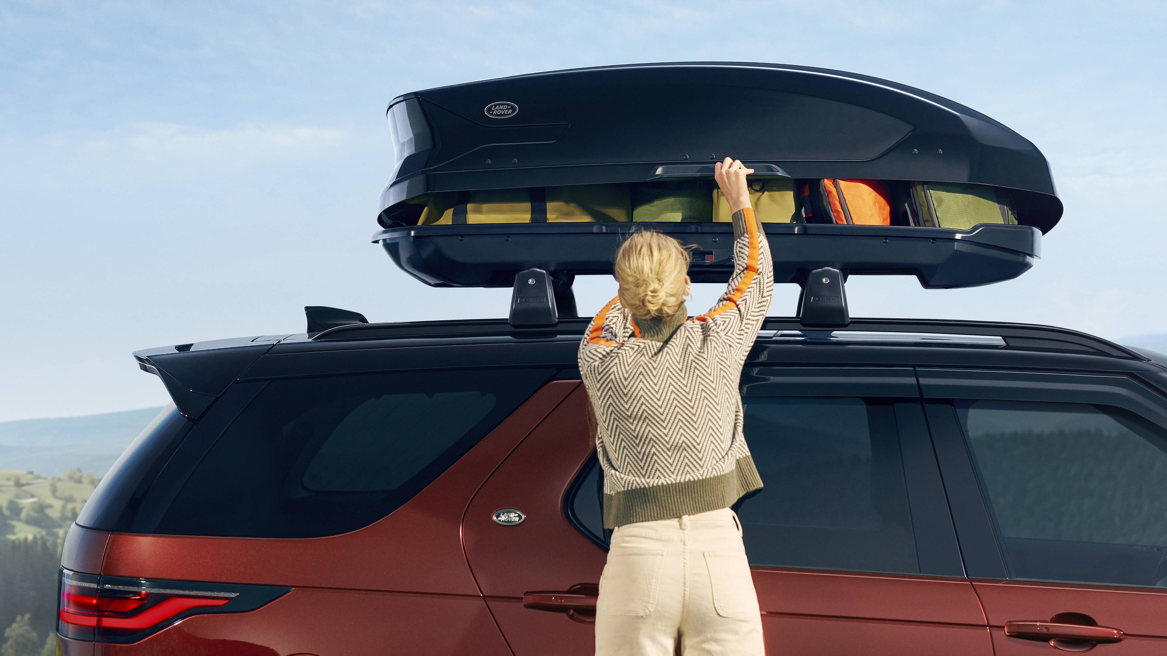 side profile of a dark red Discovery with a black roof box attached, a lady stands at the front of the image getting something out of the roof box