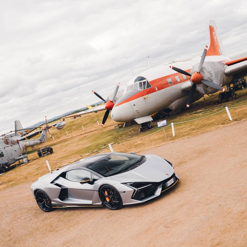 Lamborghini Revuelto in sliver with orange trims in front of a parked plane with propellers