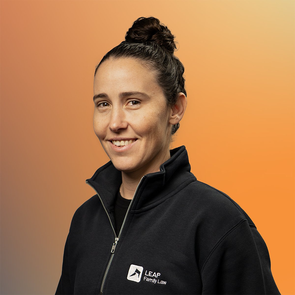A person with a bun hairstyle smiles while wearing a black zip-up jacket with "LEAP Family Law" embroidered on it, set against an orange background.