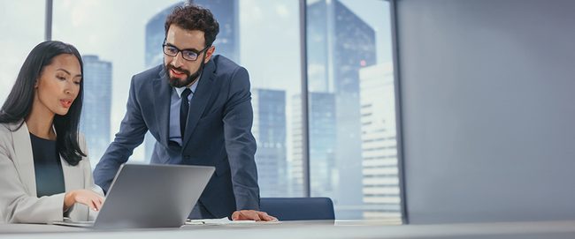 Two professionals in a modern office, one sitting and pointing at a laptop, the other standing and observing. Cityscape visible through windows.